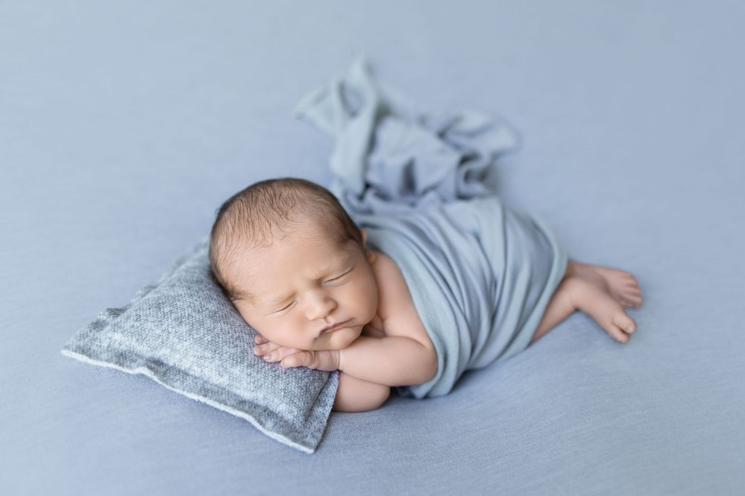 Sleeping newborn wrapped in soft gray fabric resting on textured pillow.