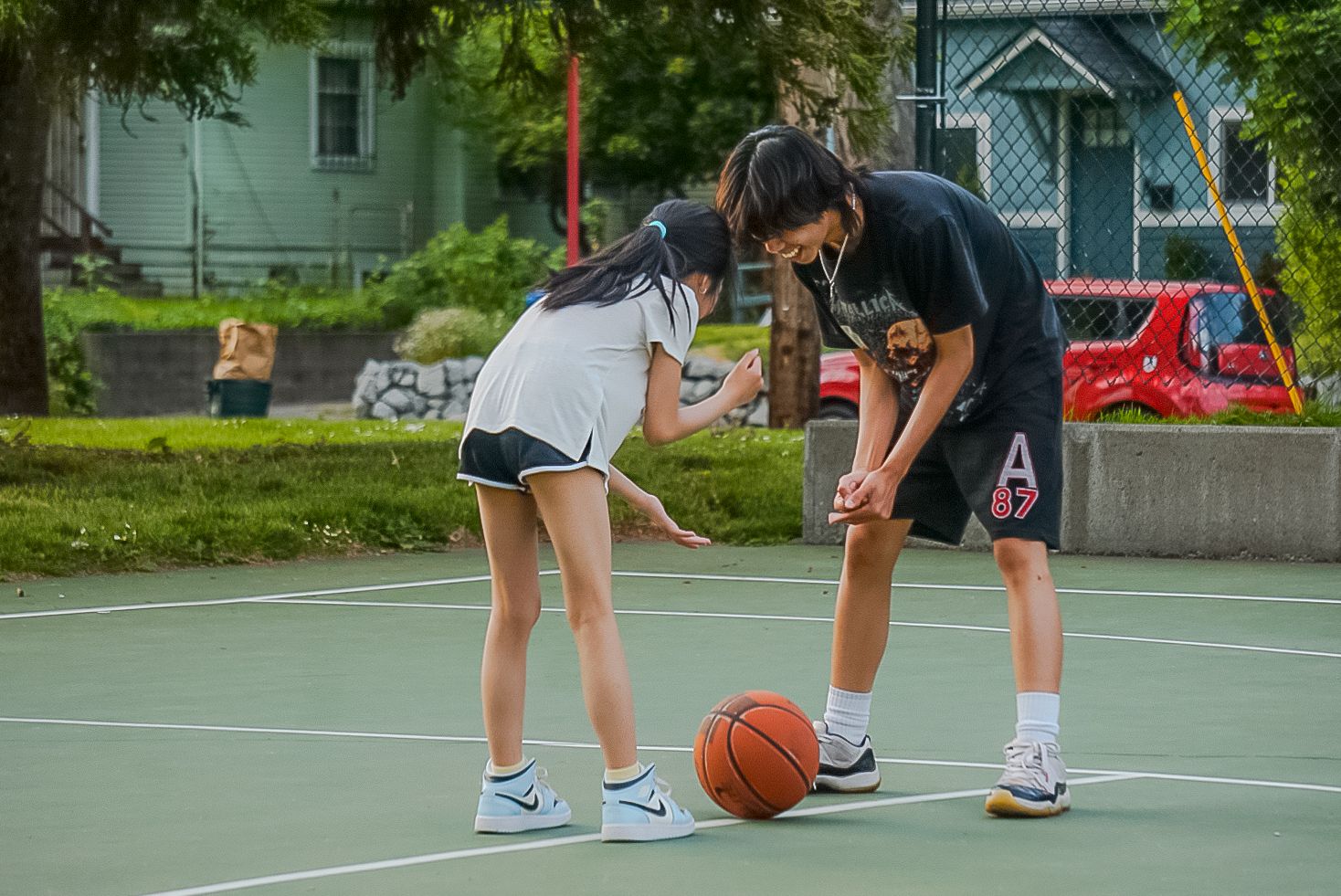 Two players practice basketball dribbling skills together on an outdoor court on a sunny day.