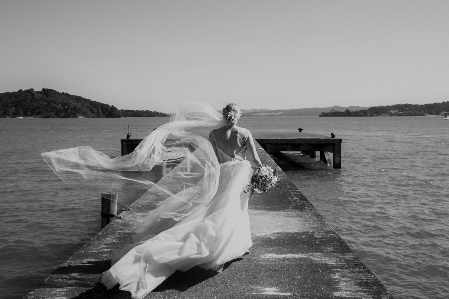 A dramatic black and white photograph of a flowing wedding dress catching the wind on a wooden pier by the ocean.