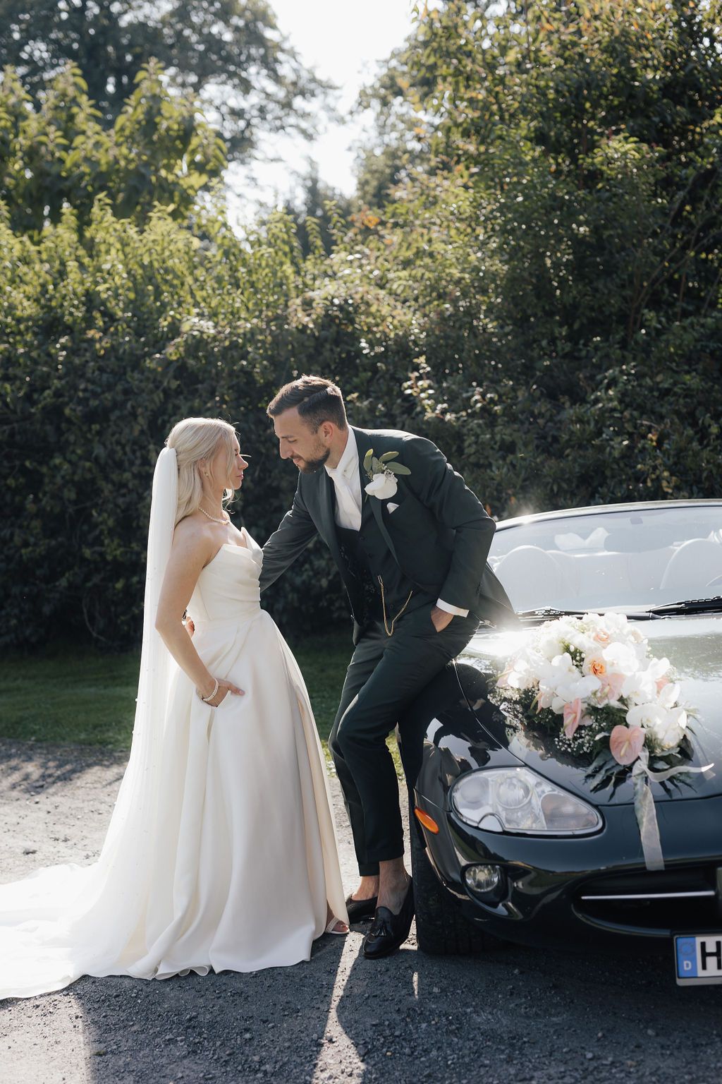Elegant bride in flowing gown poses near luxury car during wedding photoshoot.