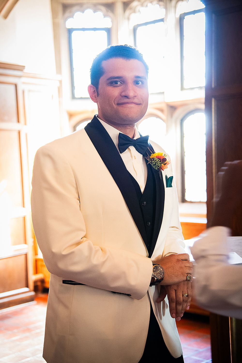 A groom poses in a white tuxedo jacket with black lapels and bowtie while wearing a small orange boutonniere. (Highland Park Rochester NY)