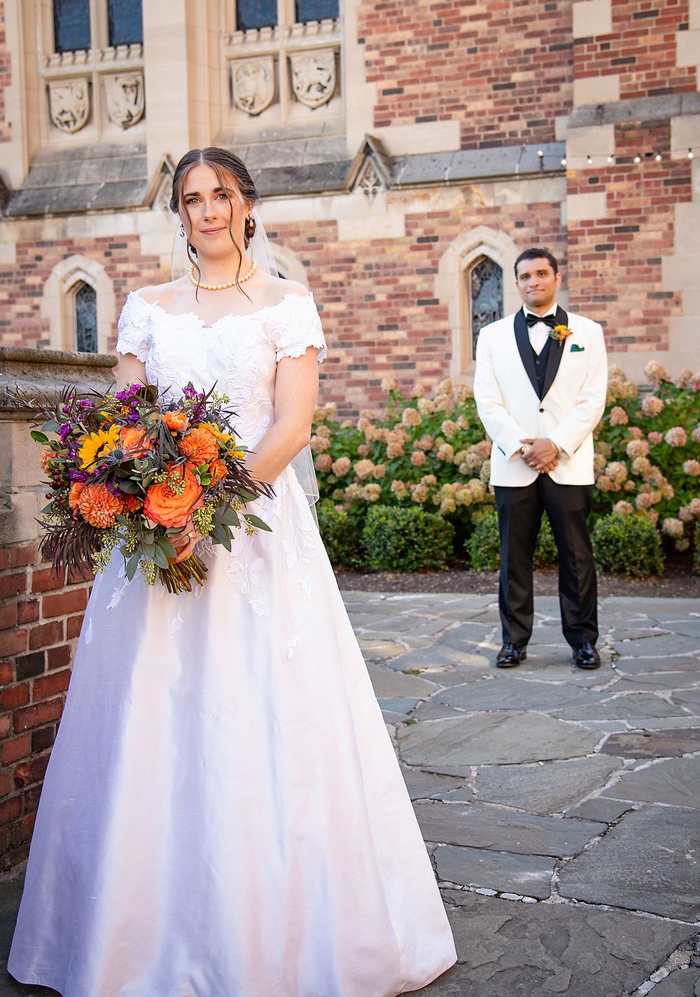 Bride in off-shoulder white gown holds vibrant autumn bouquet while groom in white tuxedo stands behind at stone church. (Highland Park Roch
