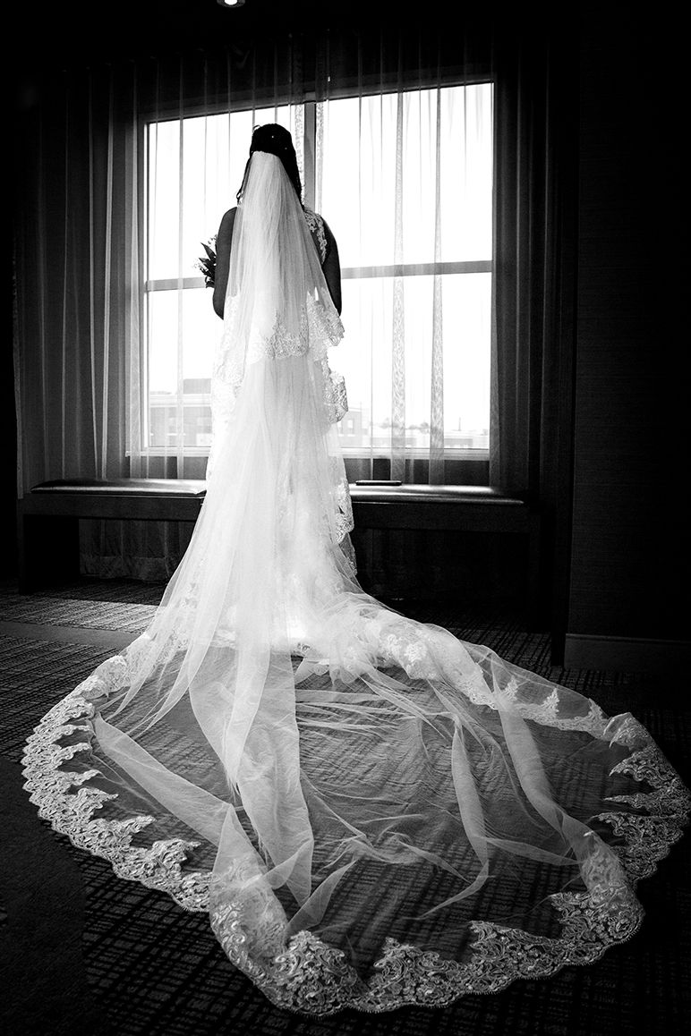 A dramatic black and white photograph of a bride in a long lace-trimmed cathedral veil silhouetted against a window.