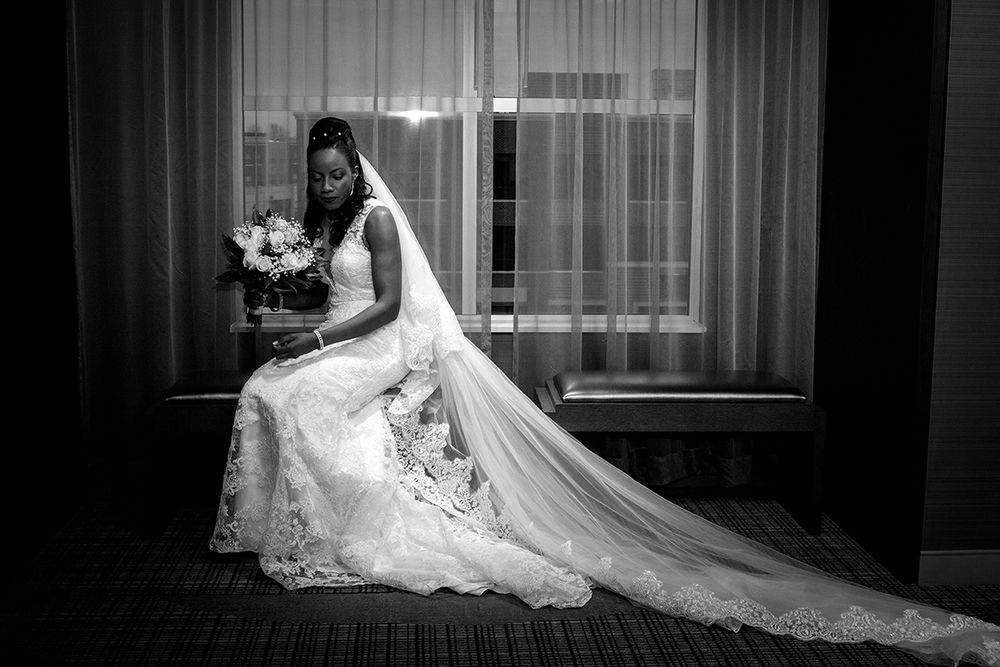 A bride in an elegant lace wedding gown with a flowing veil sits gracefully near a window in black and white photography.
