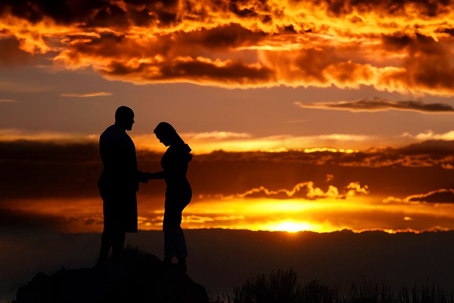 Two silhouettes stand on a hilltop against a dramatic sunset with fiery orange clouds and mountains in the background. (Henrietta New York)