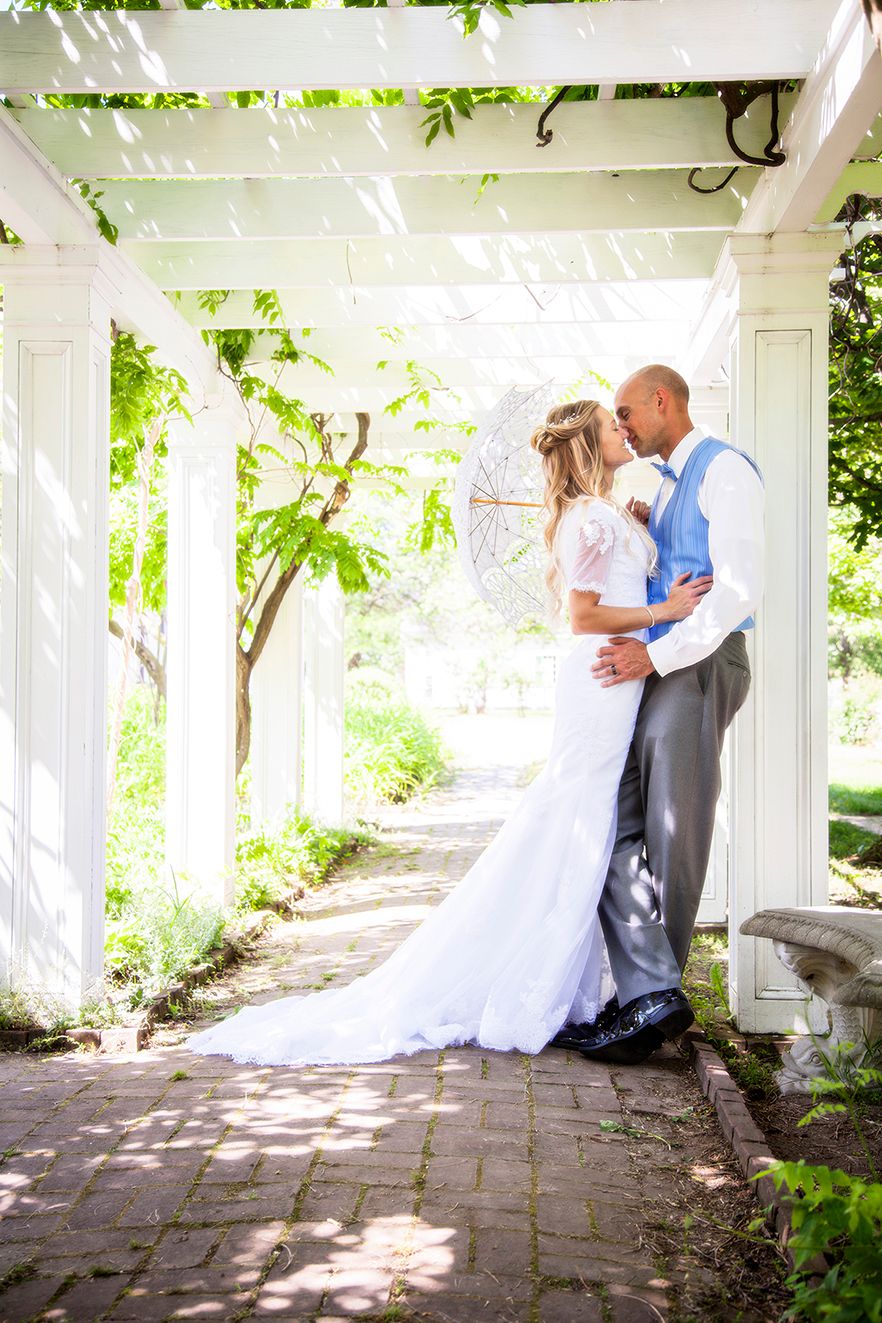 A couple shares a romantic moment under a white pergola with dappled sunlight and greenery on a stone pathway. (Brighton New York)