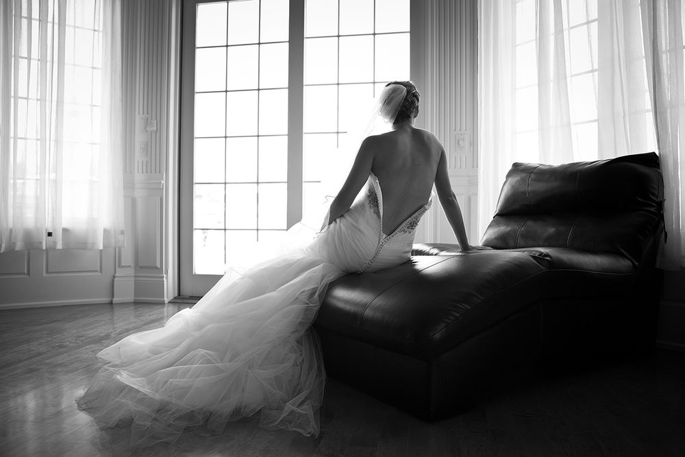 A bride in a flowing white wedding gown sits gracefully on a leather couch by sunlit windows in black and white.