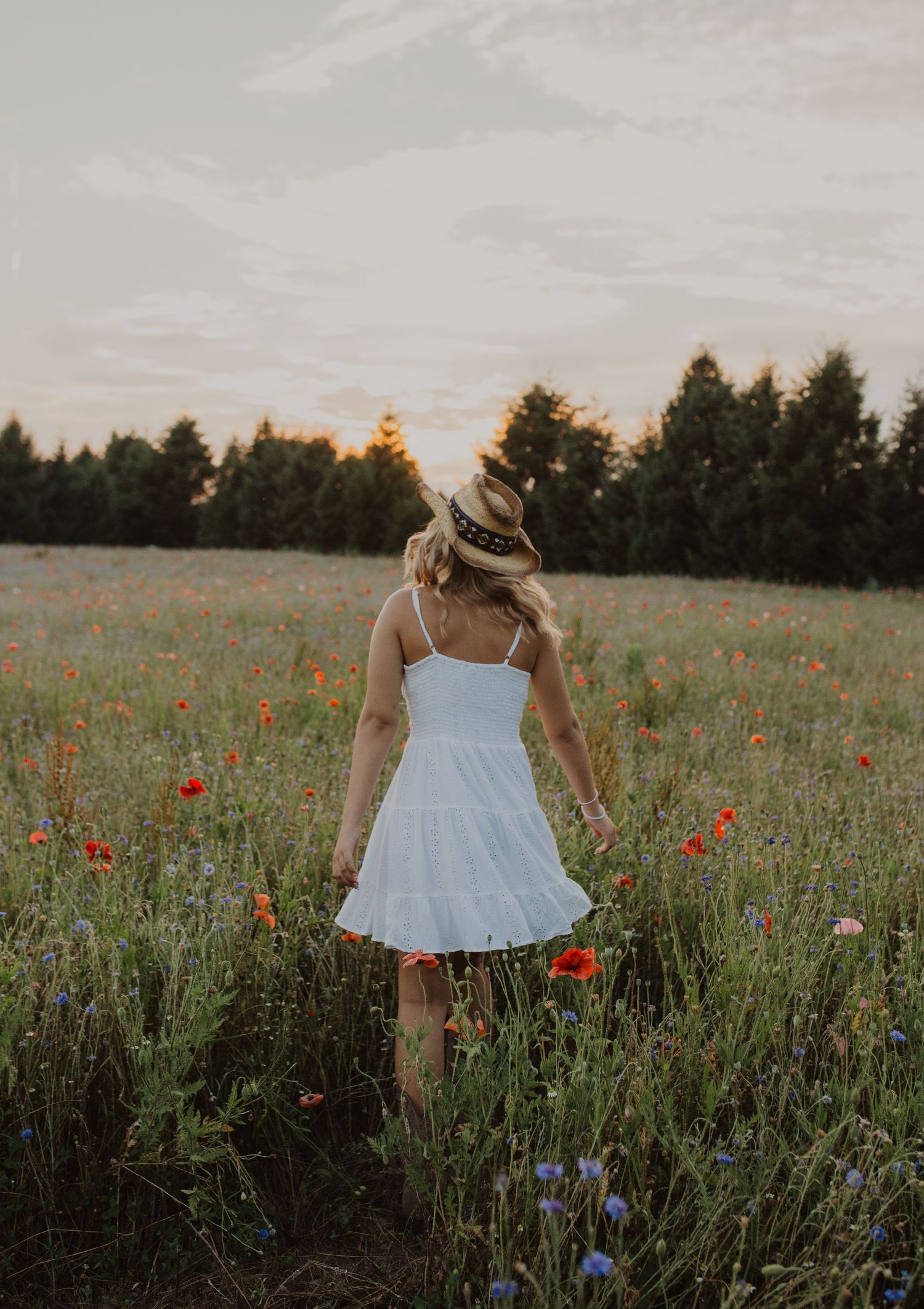 Figure in flowing white dress walks through field of red poppies and blue wildflowers.
