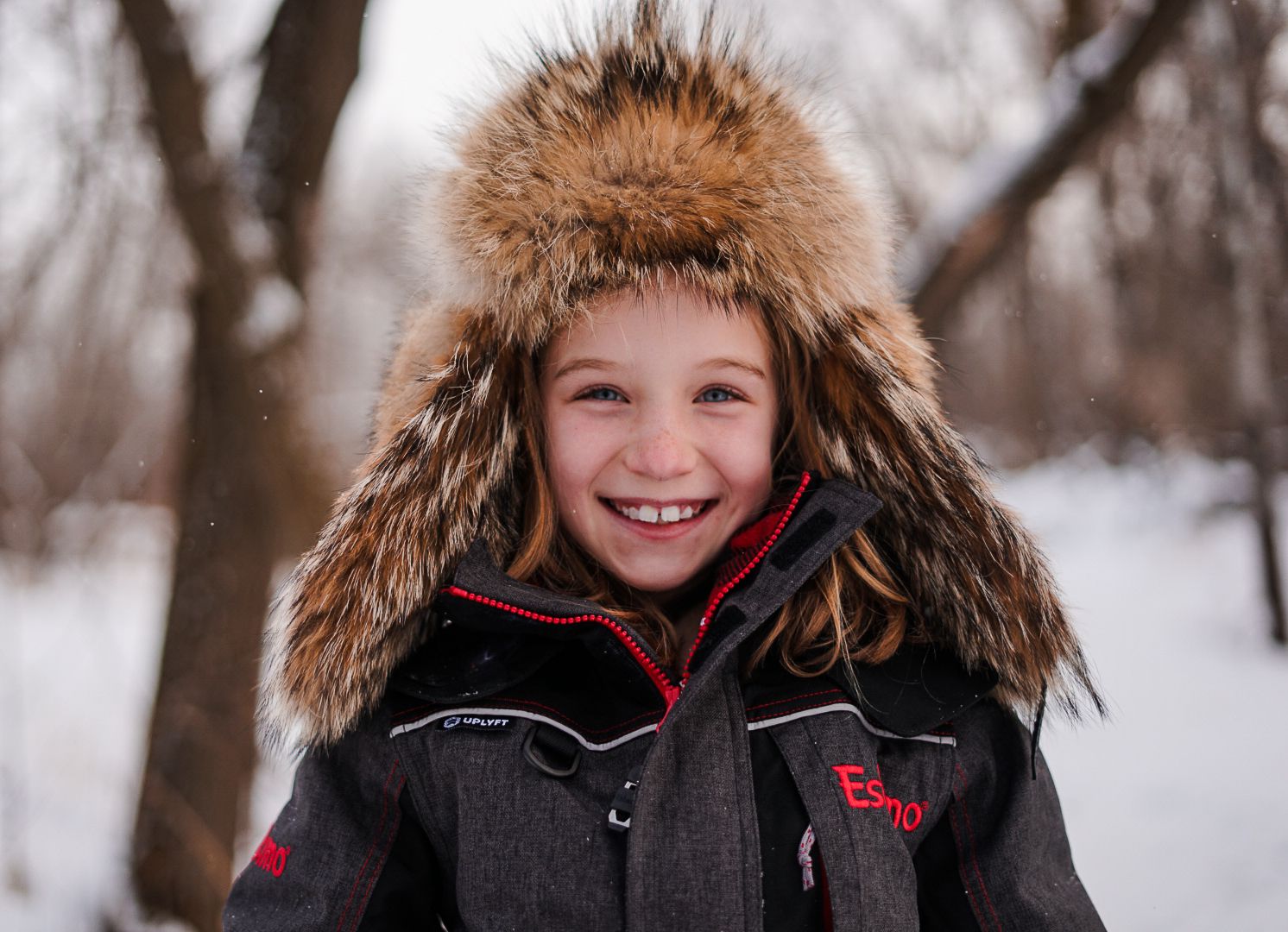 Child wearing furry winter hat smiling outdoors in snowy weather.