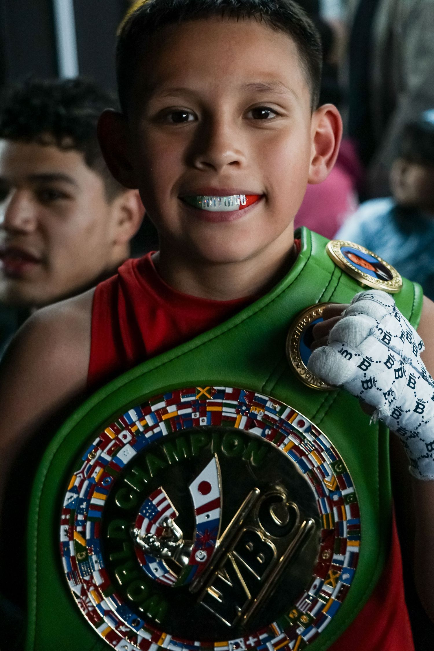 Young boxing champion smiles while displaying a green championship belt decorated with international flags at a competition.