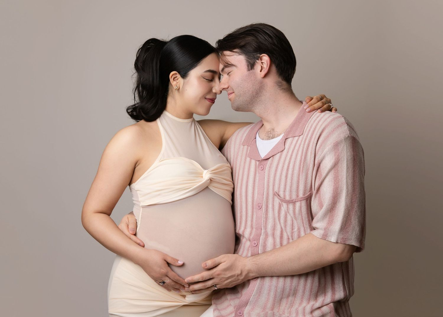 A couple in soft pastel clothing shares an intimate moment while expecting a baby, set against a neutral backdrop.