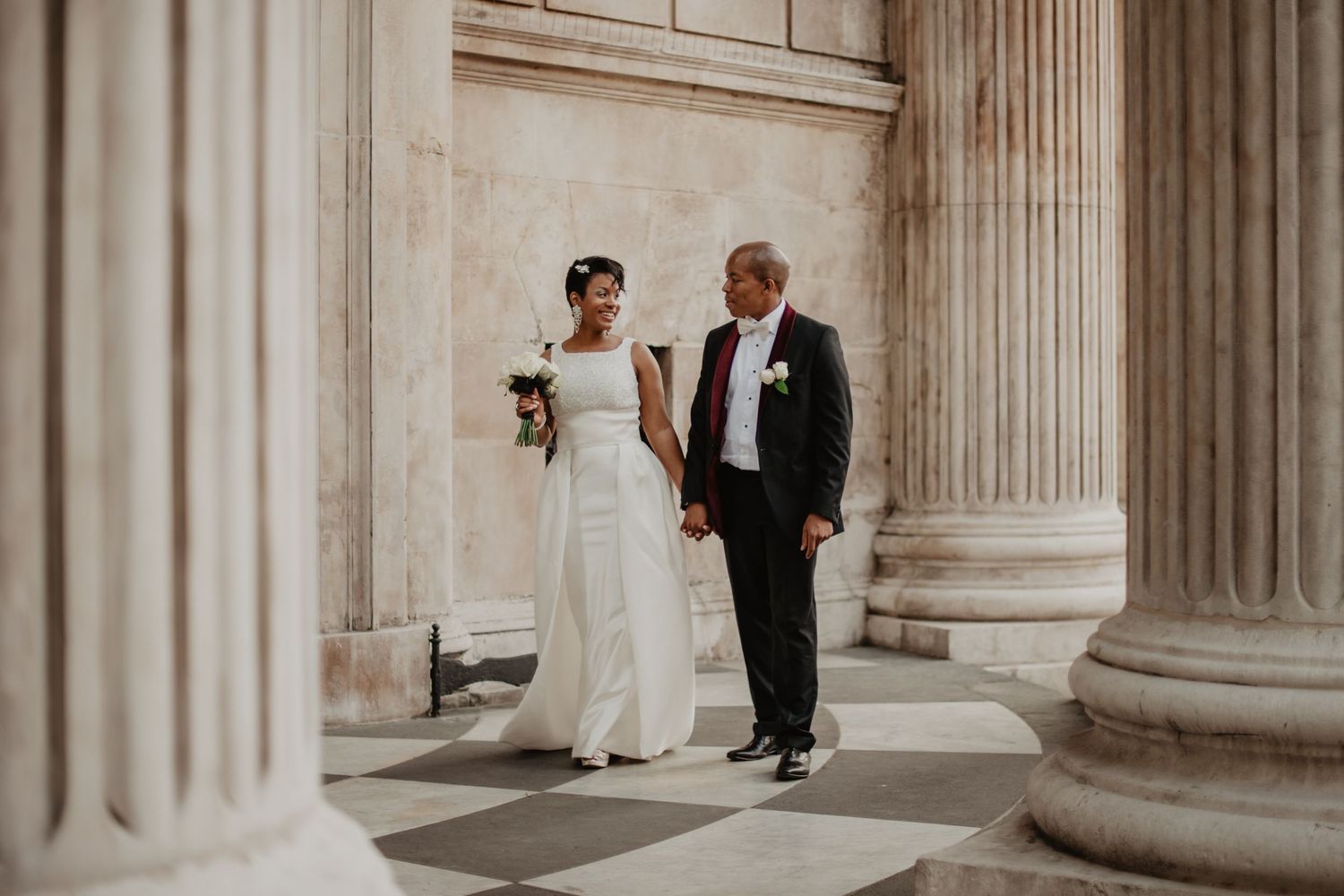 Bride and groom walk holding hands between imposing columns at St Paul's Cathedral. Bride in white strapless gown, groom in black suit