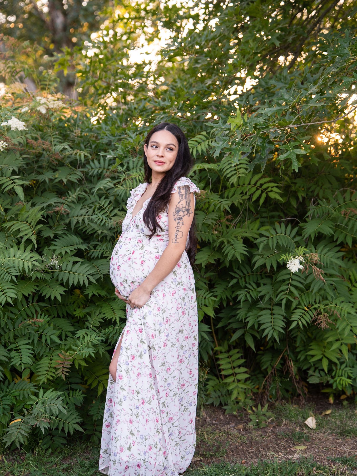 A person in a floral maxi dress stands outdoors among lush green foliage during golden hour sunlight.