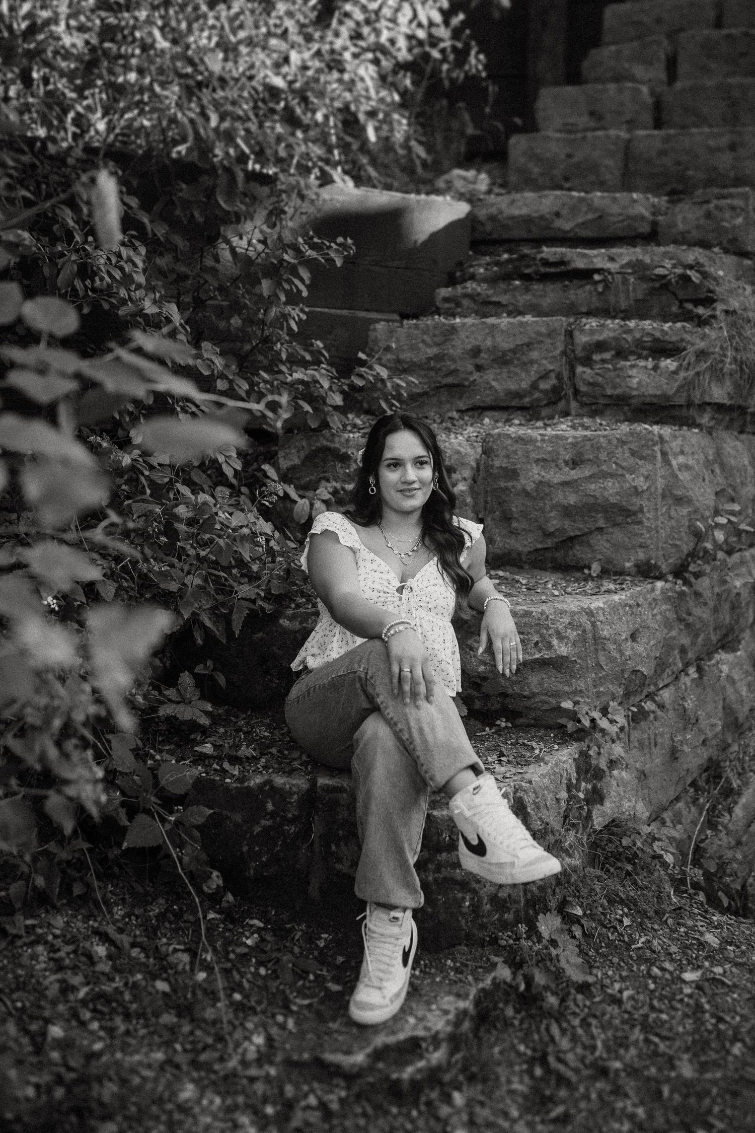 Black and white portrait of someone sitting casually on stone steps surrounded by foliage wearing white sneakers and casual attire.