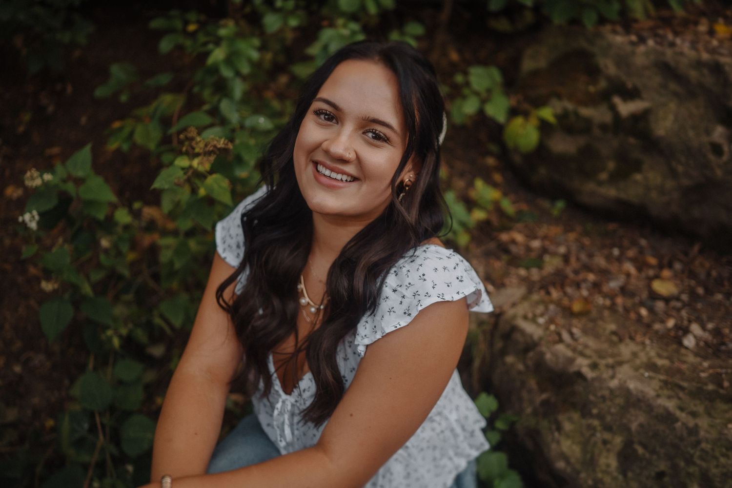 Seated against stone wall with autumn foliage, someone wearing white lace dress smiles warmly in outdoor portrait setting.