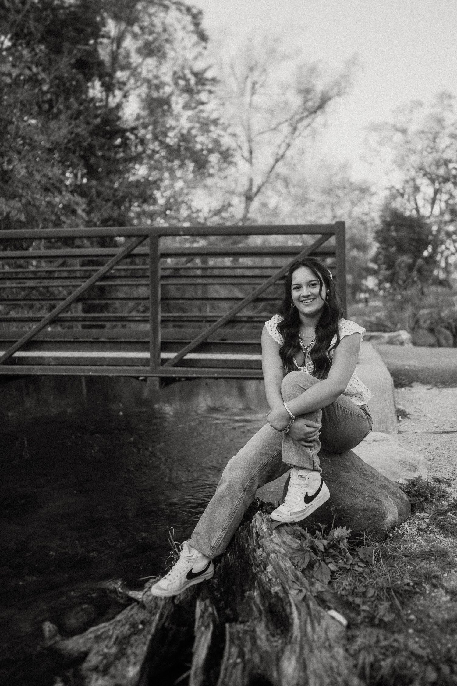 Black and white portrait of someone sitting on a log by a bridge, smiling outdoors in a natural setting.