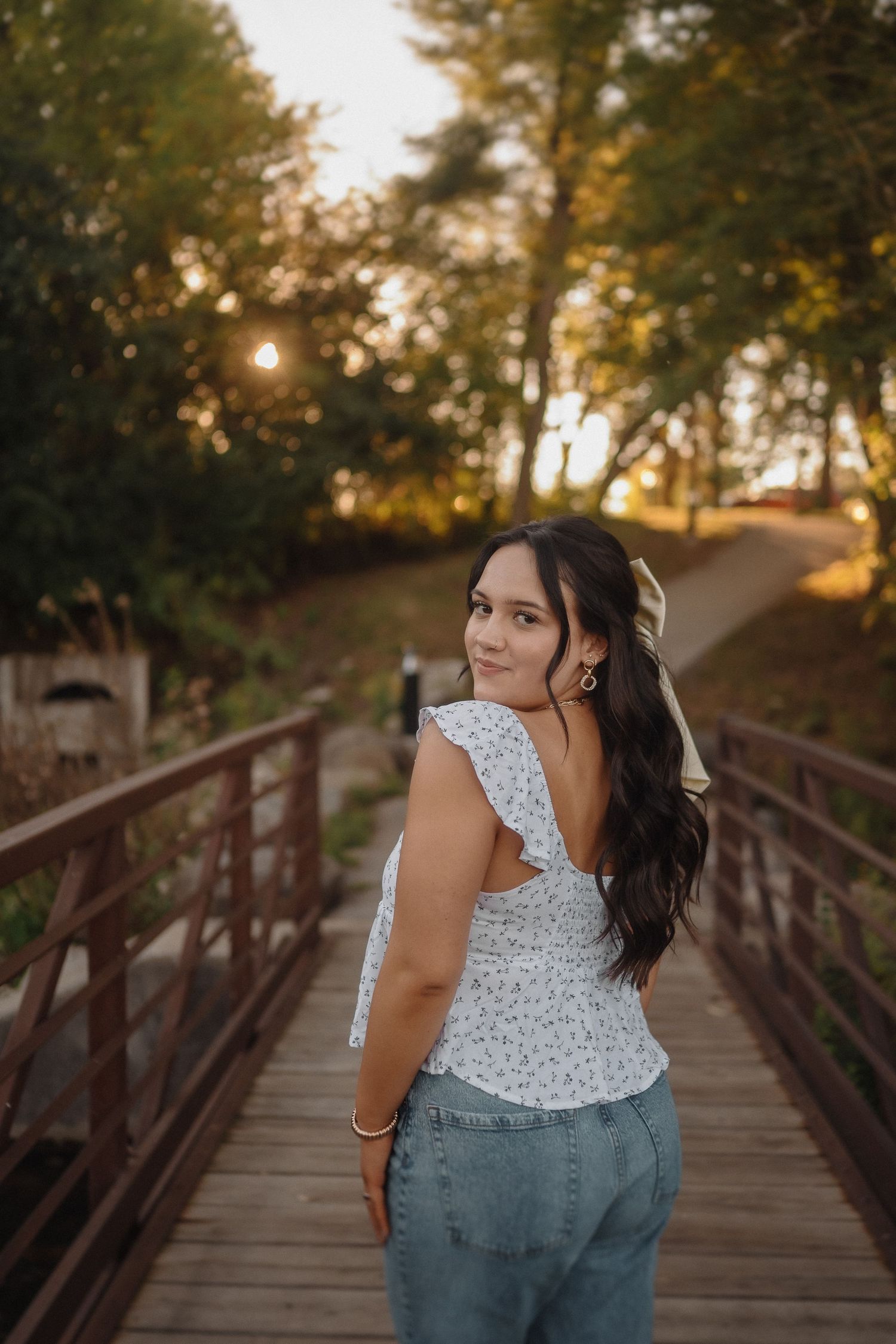 Woman in floral top poses on wooden bridge during sunset surrounded by autumn trees.