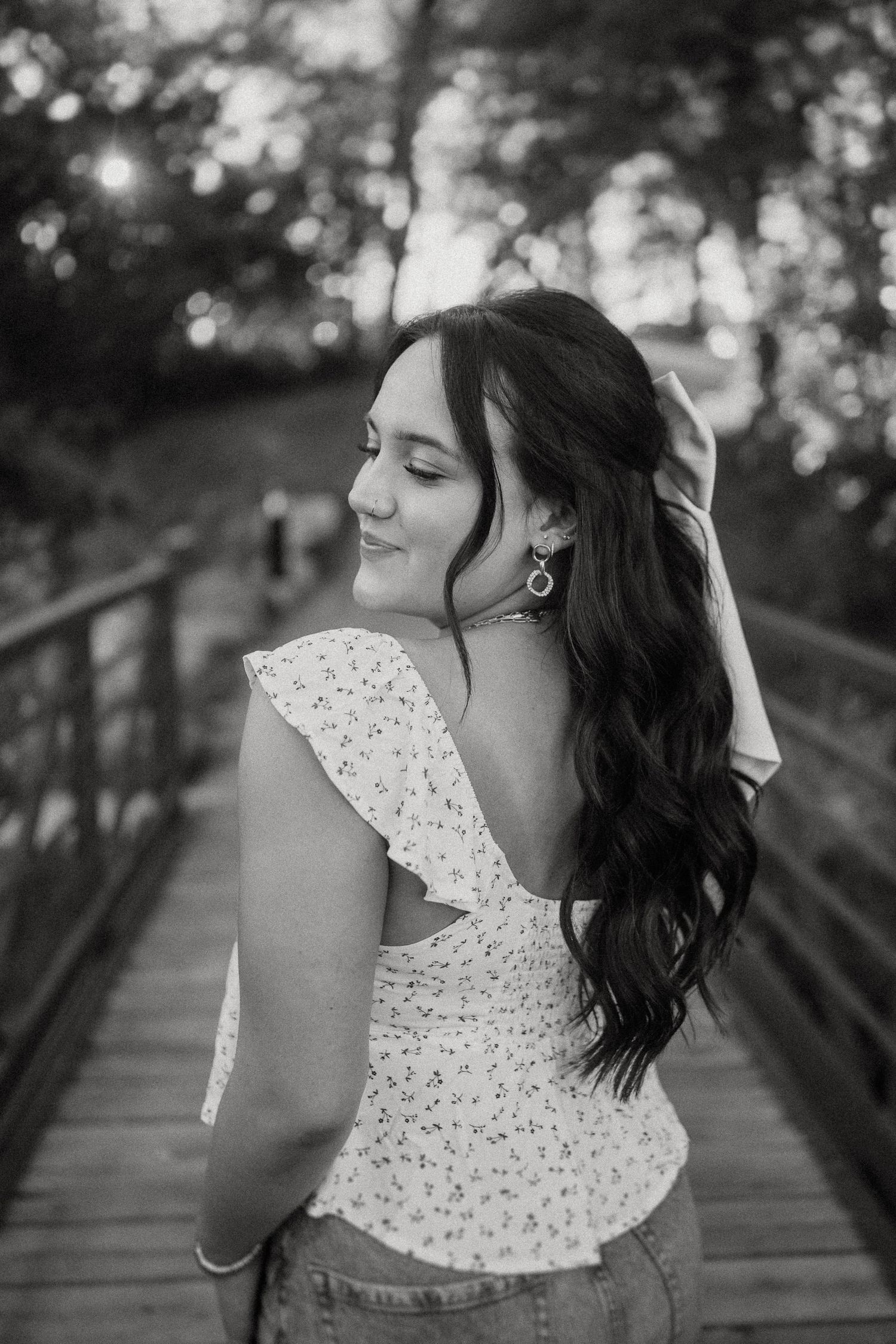 Black and white portrait of woman in lace top on wooden bridge with bokeh trees in background.