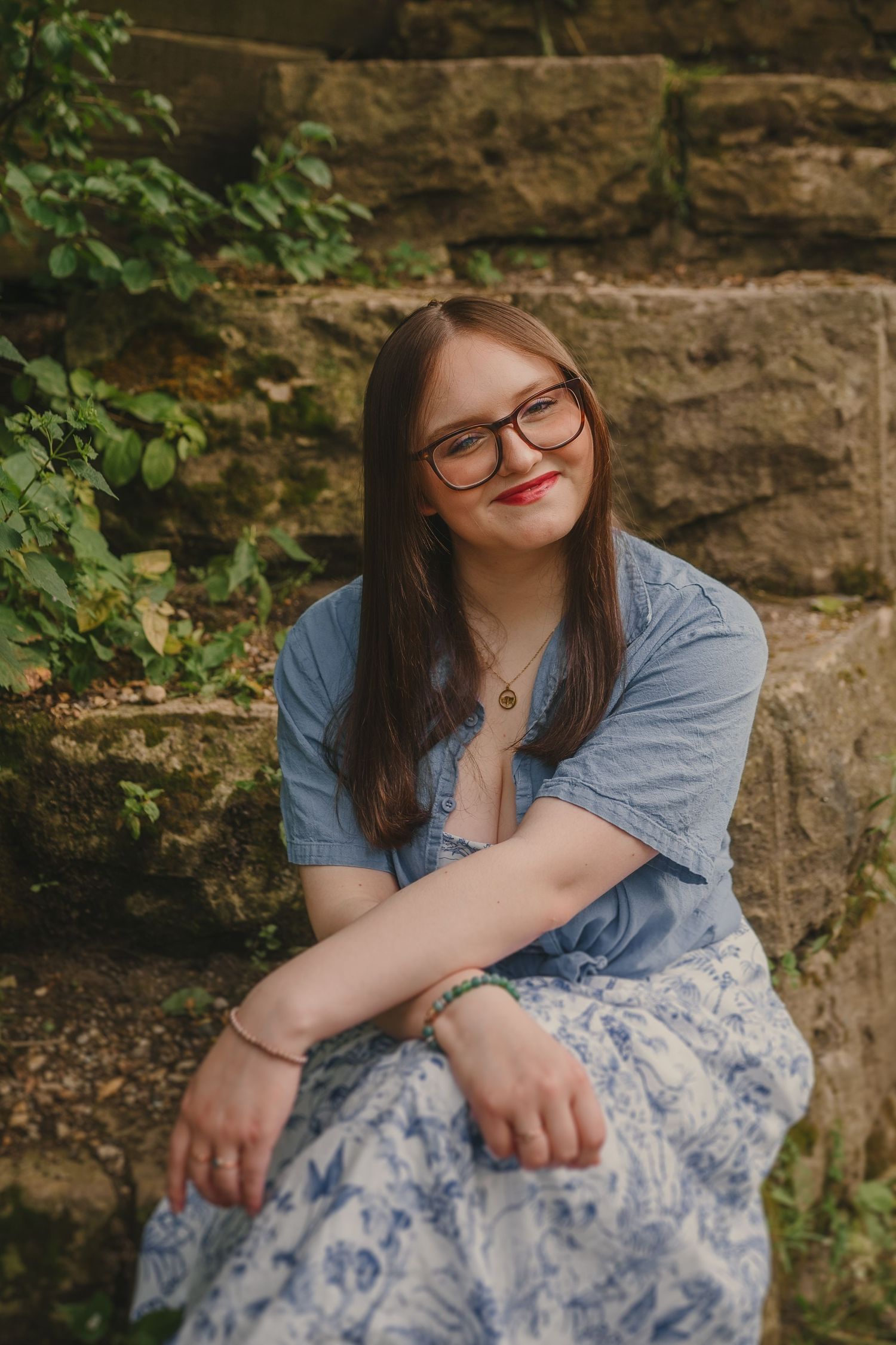 A portrait of someone in glasses and denim sitting against a stone wall with greenery in a floral dress.