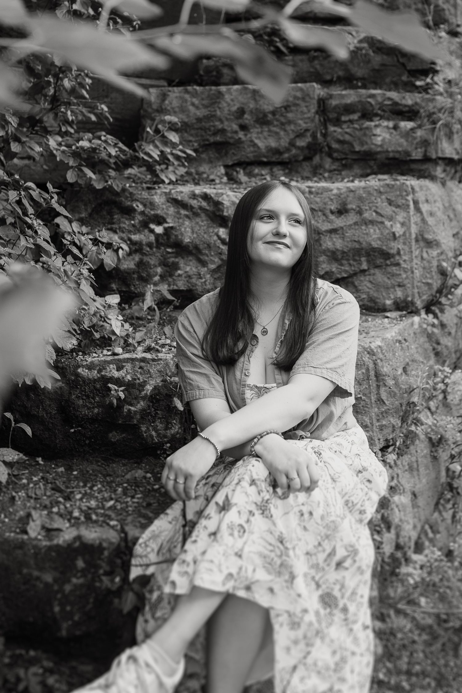 Black and white portrait of someone sitting outdoors against a stone wall wearing a floral dress and long dark hair.
