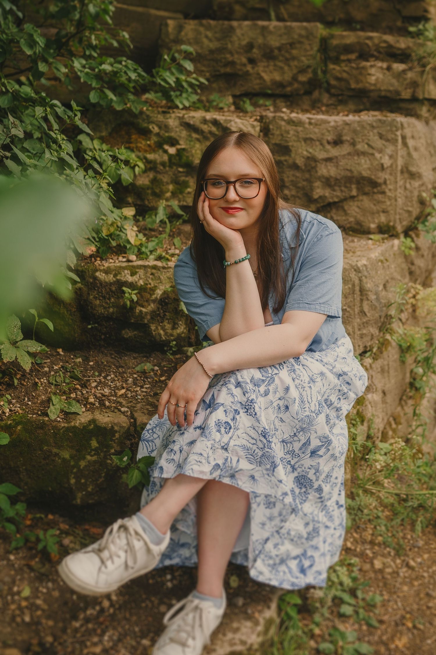 A portrait in a blue shirt and floral dress sitting on stone steps surrounded by greenery.