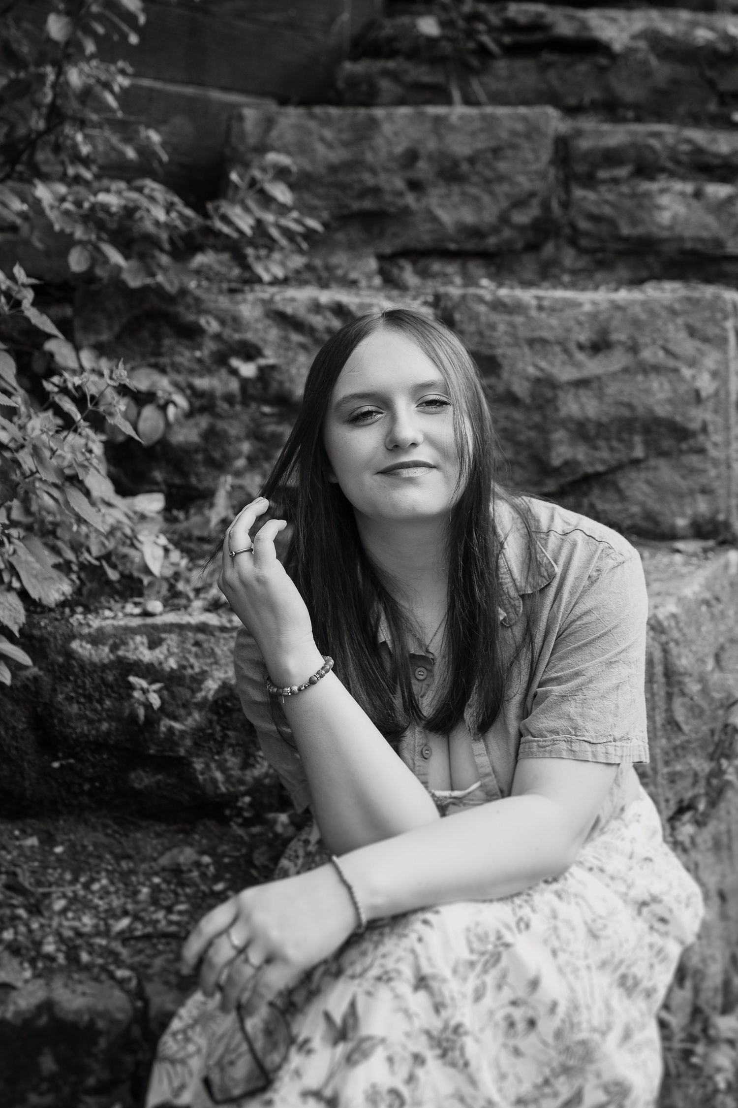 Black and white portrait of a relaxed young woman in a patterned skirt sitting by a stone wall outdoors.