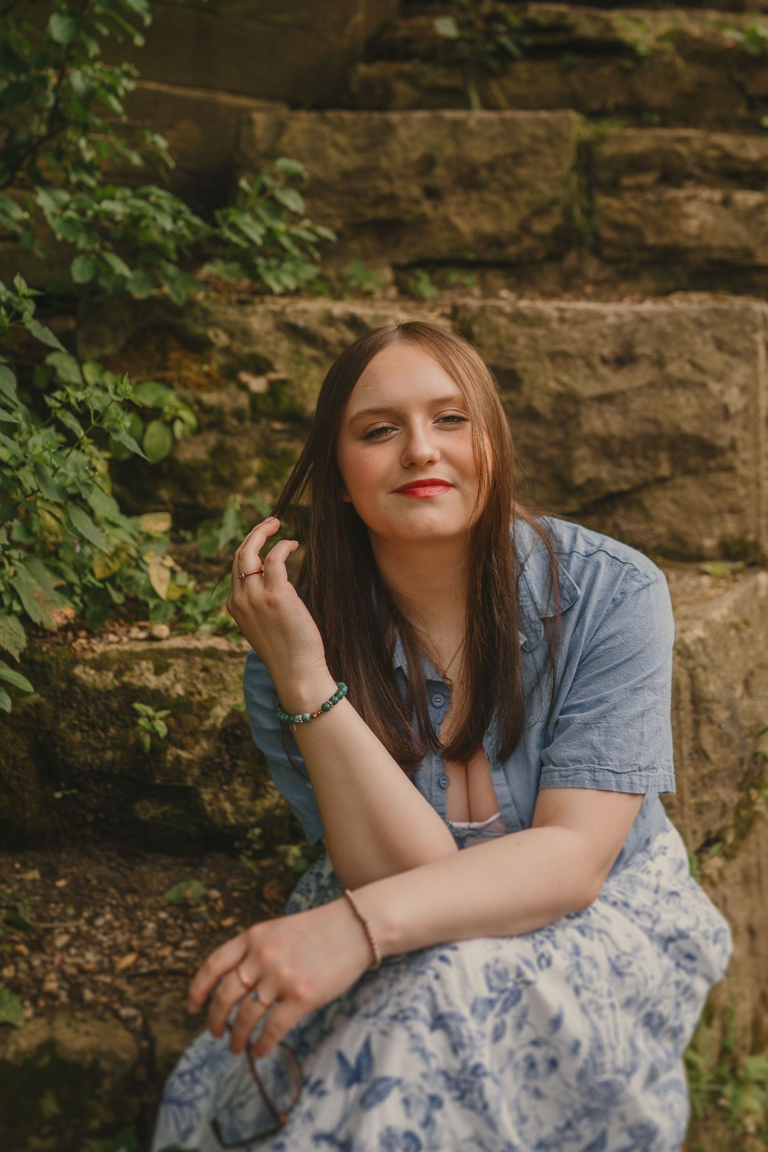Portrait of someone in a chambray shirt and floral skirt sitting by a stone wall with green vines.
