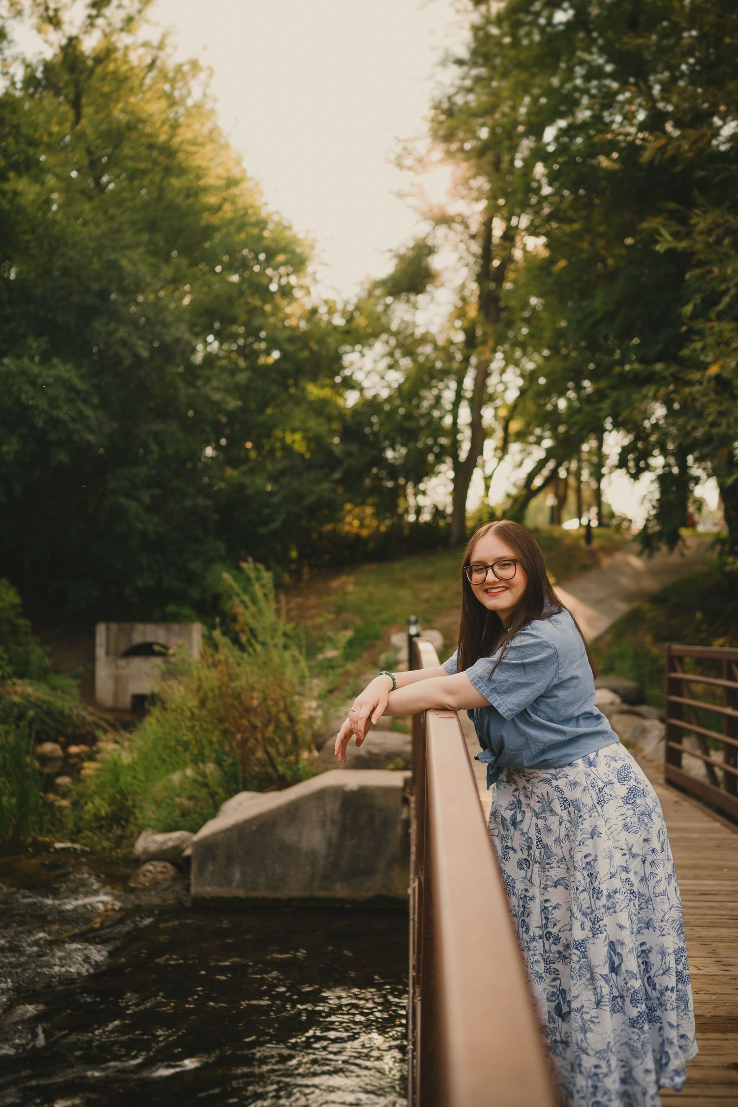 Person in denim jacket and floral skirt stands on wooden bridge overlooking stream in wooded park setting.