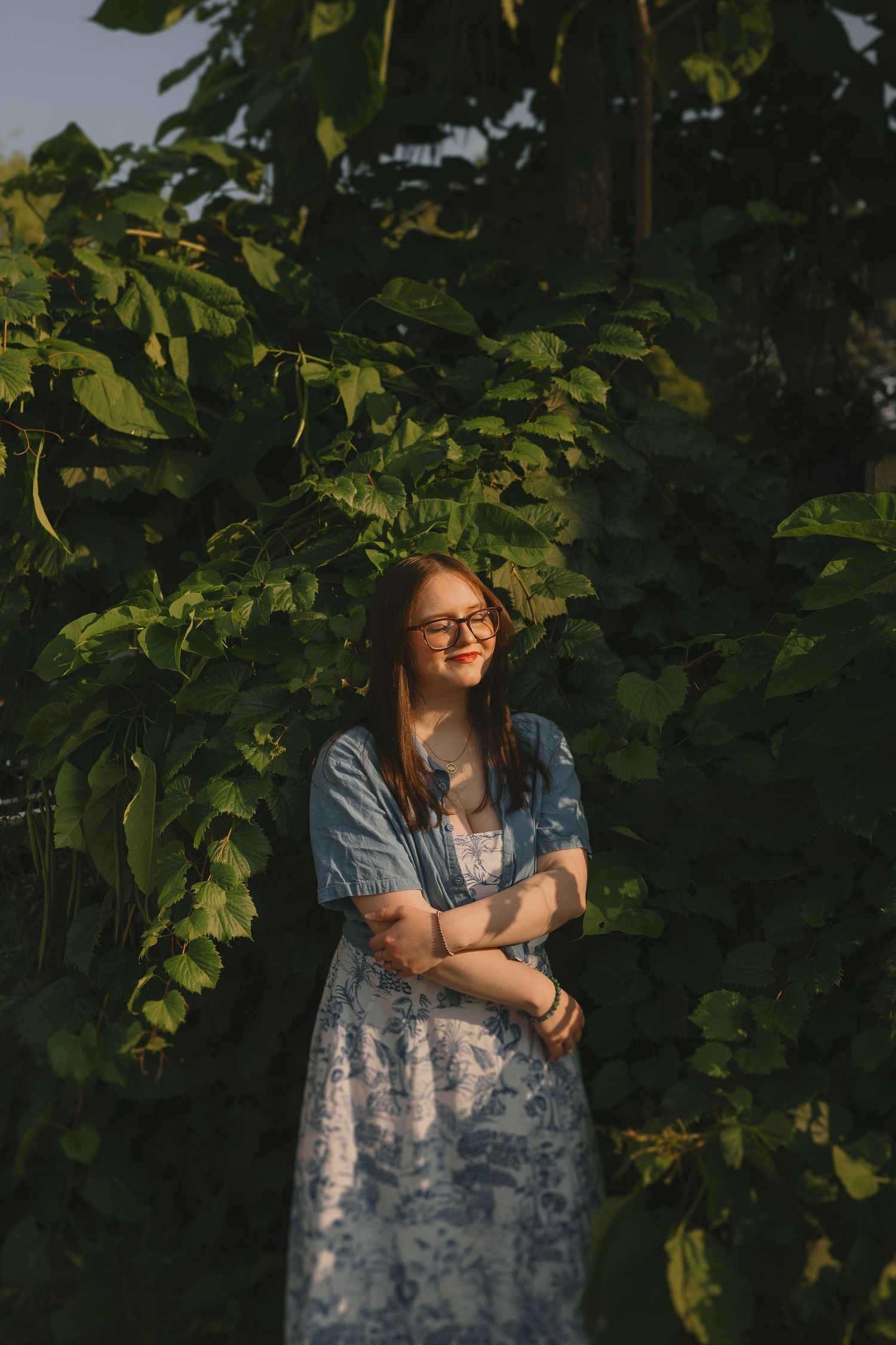 A figure in a blue denim dress stands against lush green foliage in warm sunlight.