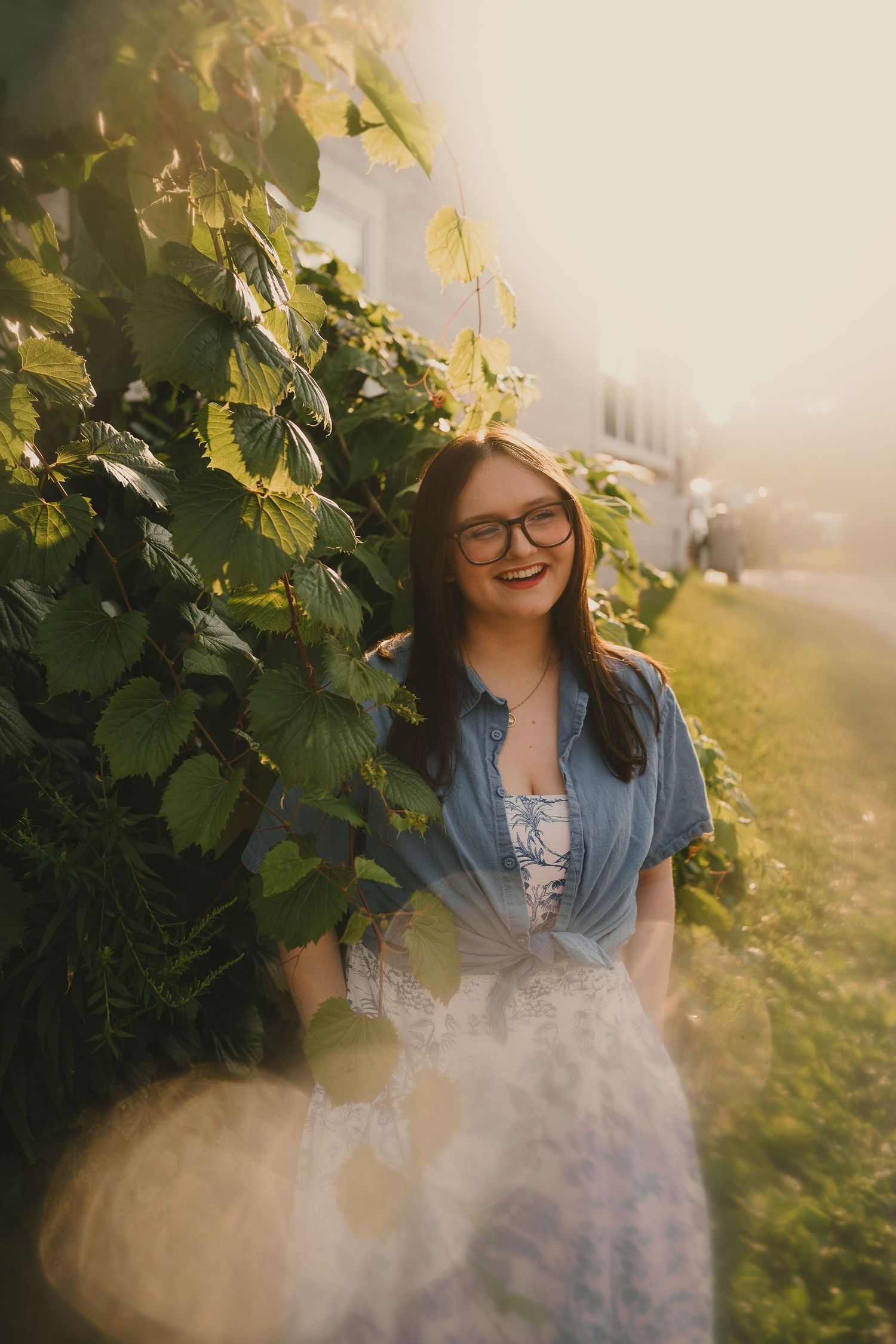 Portrait by green vines wearing a blue top and floral dress in warm sunlight.