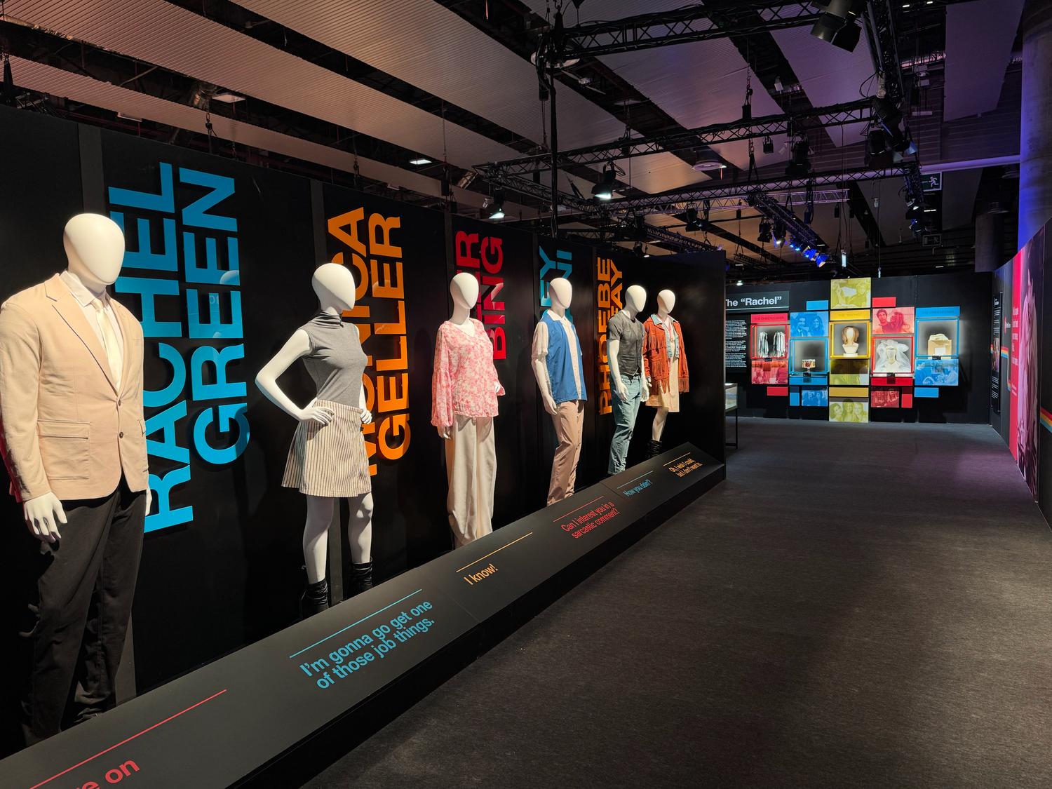 Exhibition display featuring mannequins in retro clothing with neon text graphics on black walls.