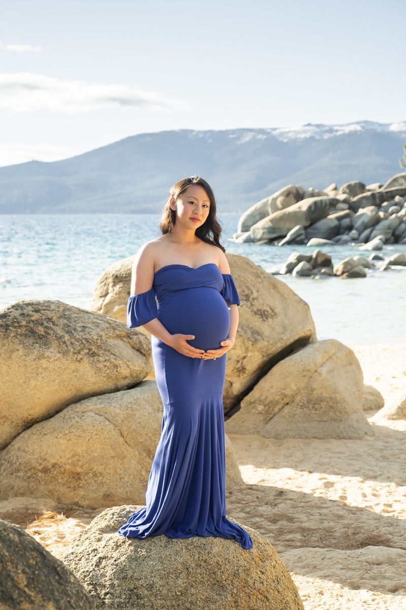 A pregnant woman in a blue off-shoulder gown poses gracefully on large rocks by a scenic mountain lake.