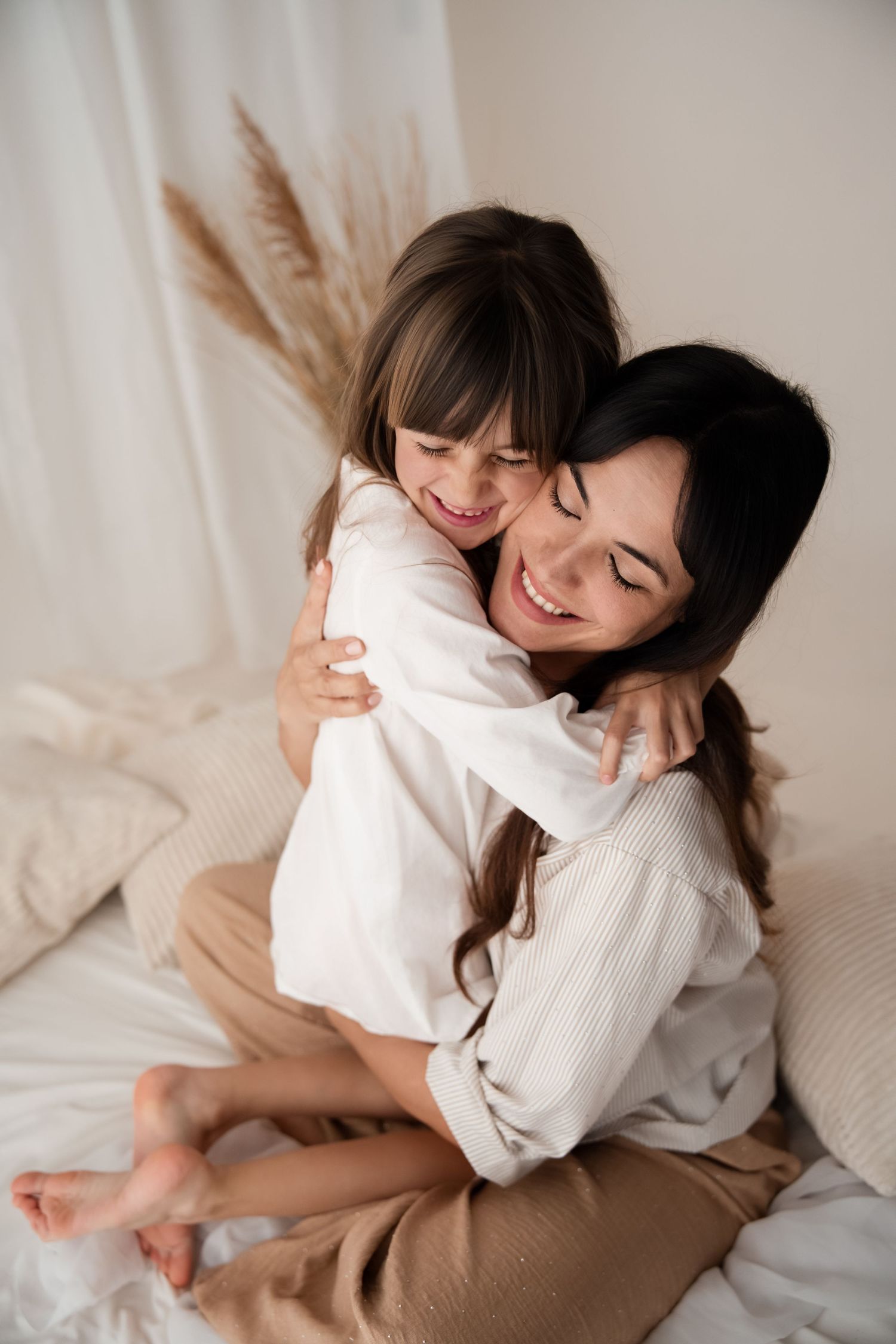 A dark-haired mother and daughter share a warm embrace while sitting on a cozy bed.