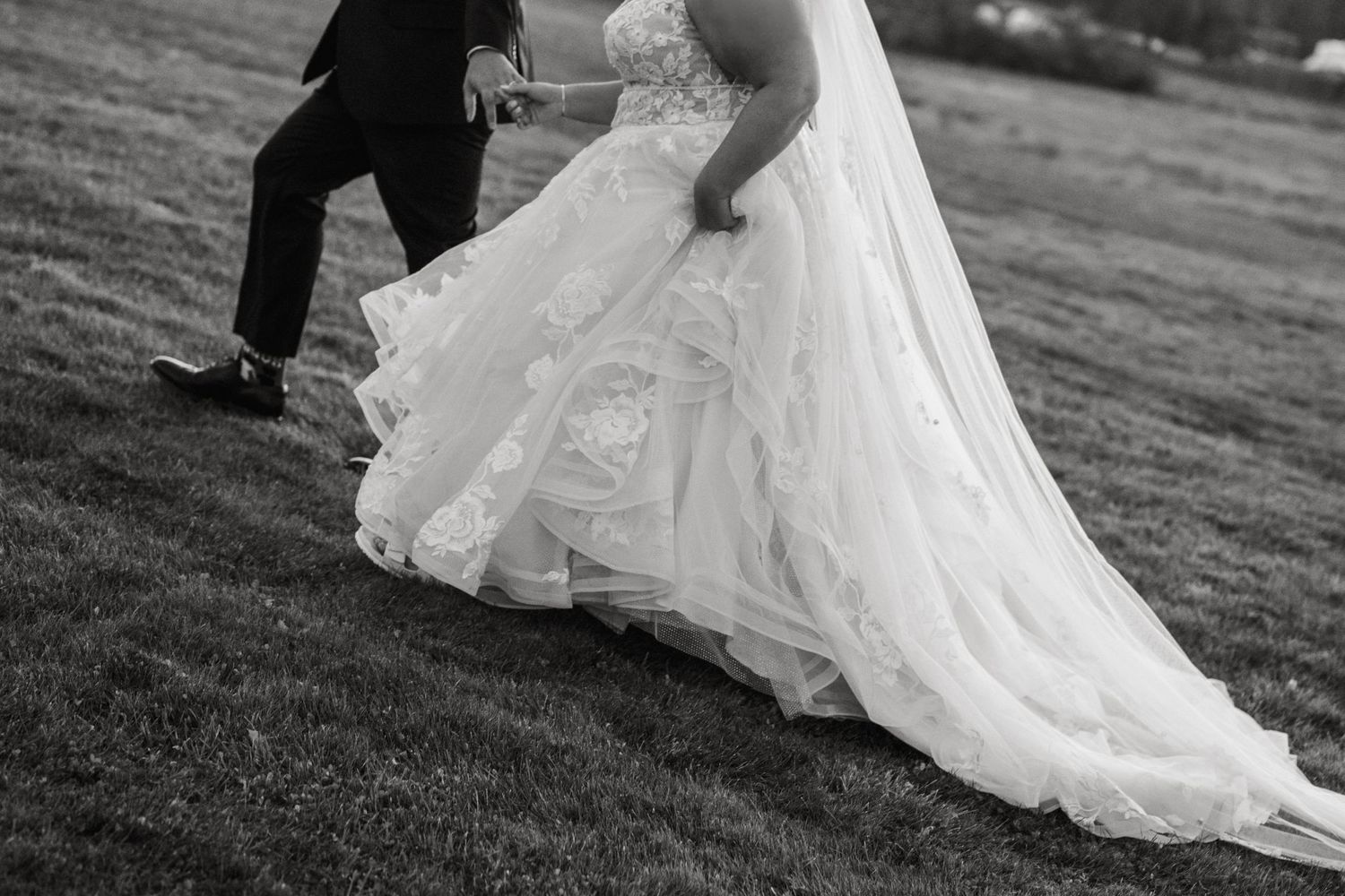 Black and white photo of a flowing white wedding dress train trailing on grass with formal shoes visible.