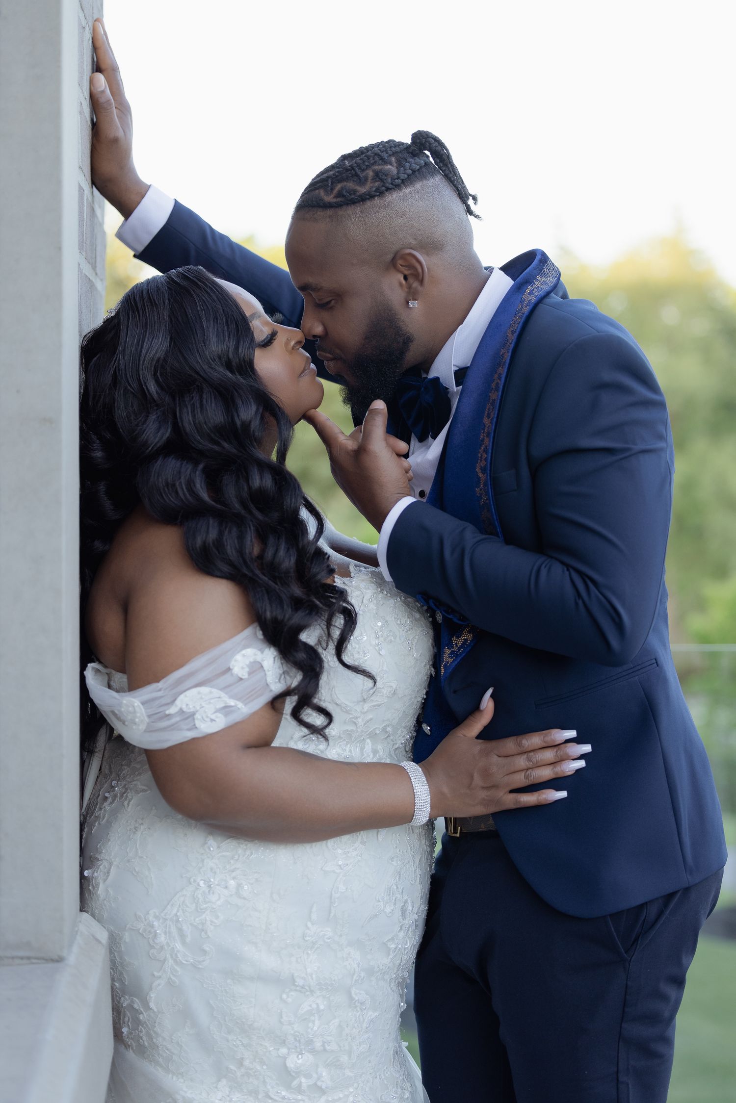 A romantic wedding photo showcases a couple in formal attire sharing an intimate moment against a stone pillar outdoors.