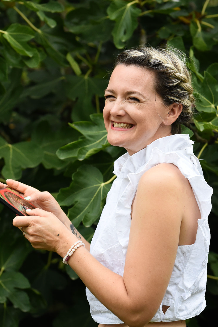 A person wearing a white sleeveless top smiles while holding a mobile phone against a green leafy background.