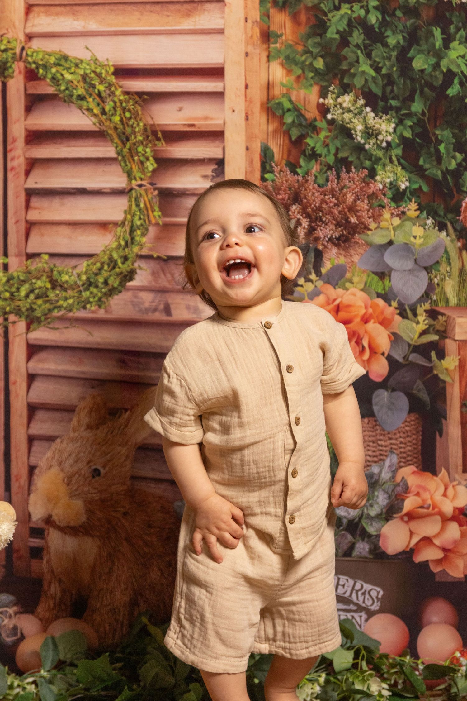 Adorable baby in beige outfit laughs and plays during a rustic photo session with autumn decorations.