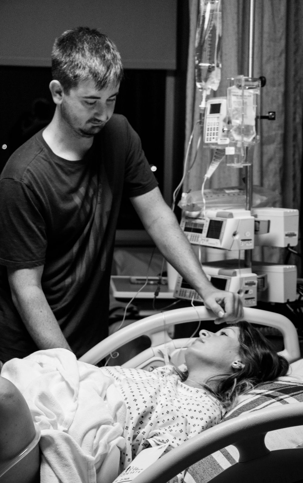 Black and white photo of person receiving medical care in hospital bed with IV equipment visible in background.