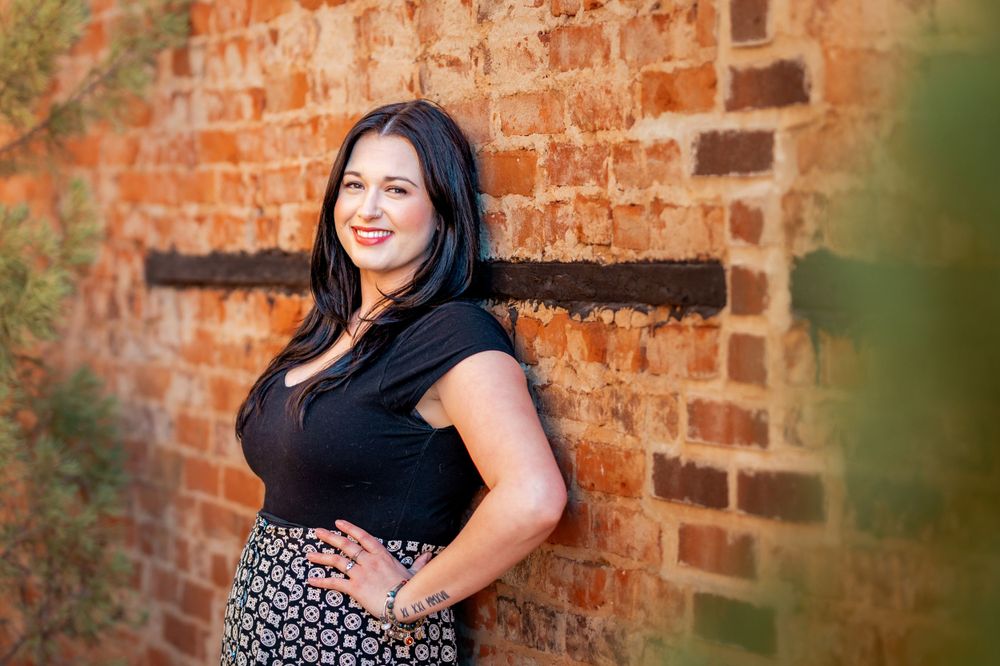 Happy figure in black top poses against rustic brick wall in outdoor portrait setting.