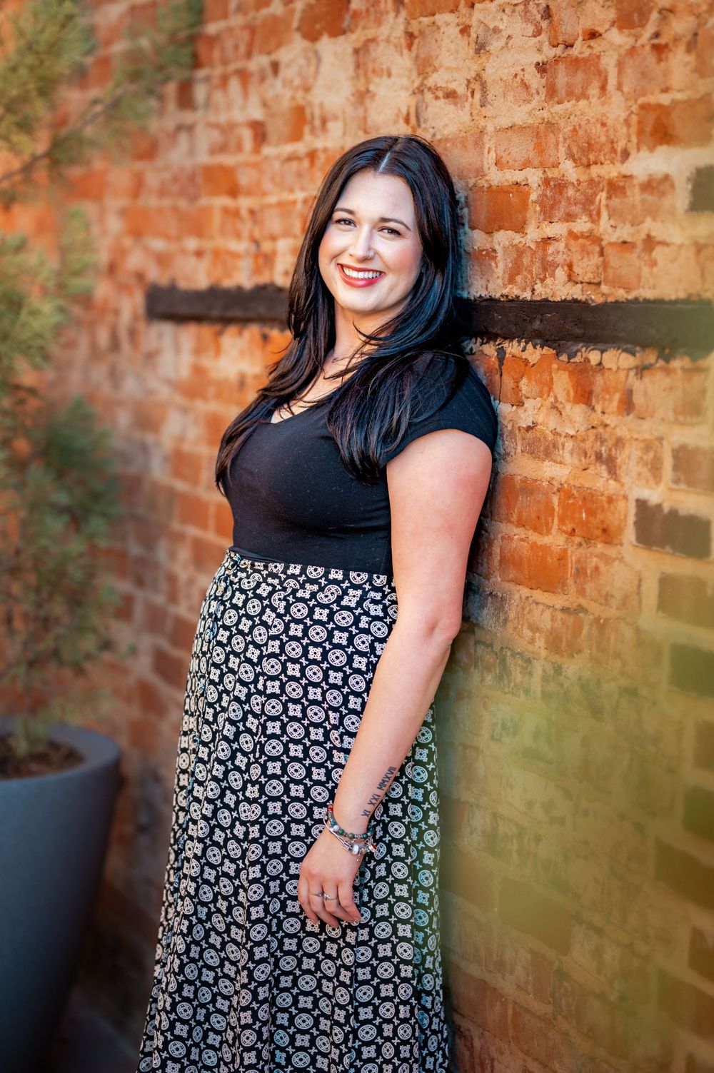 A woman in a black top and patterned maxi skirt stands smiling against a rustic brick wall outdoors.