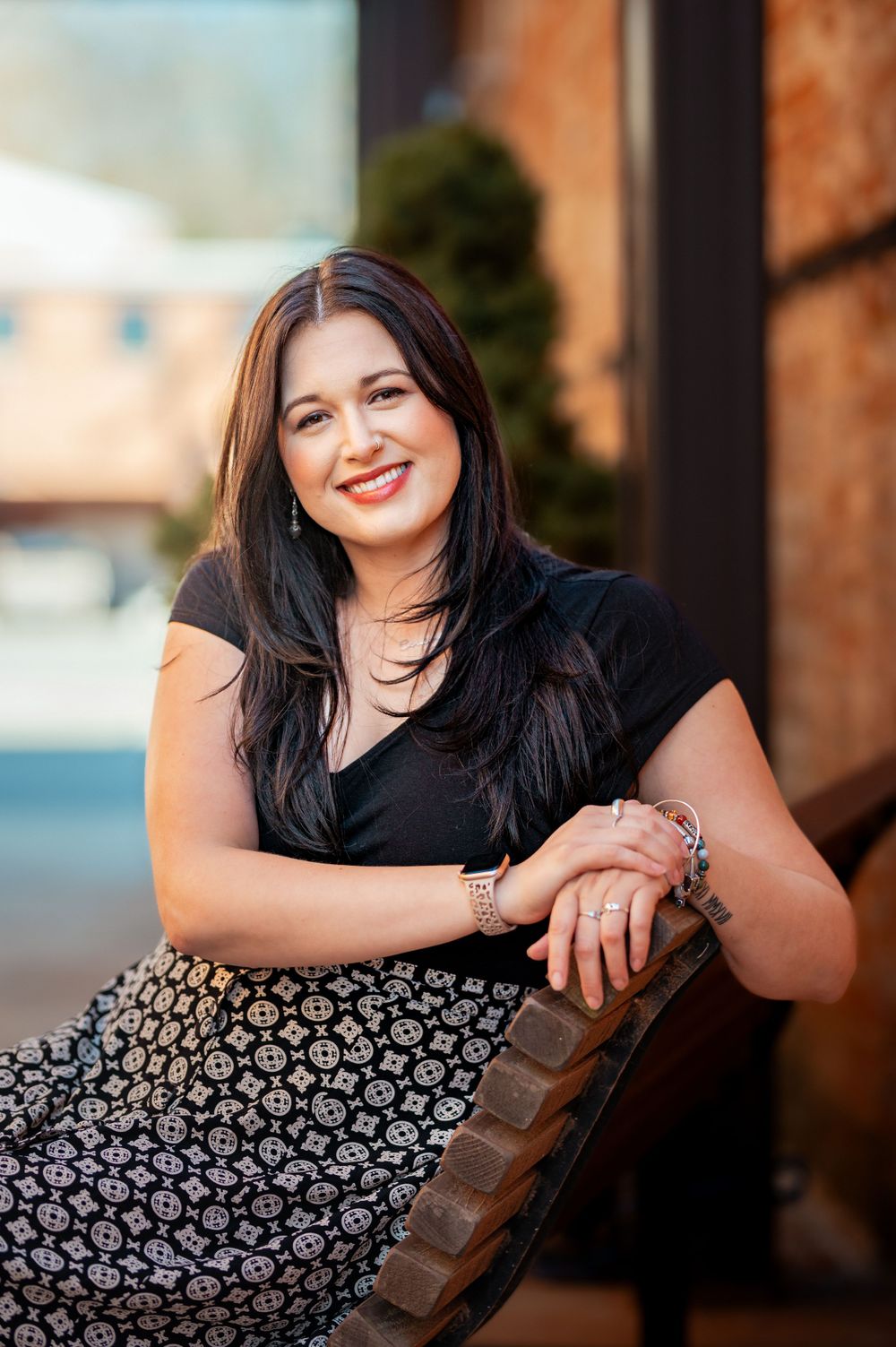Woman in black top and patterned skirt sits casually outdoors with a warm smile against brick wall background.