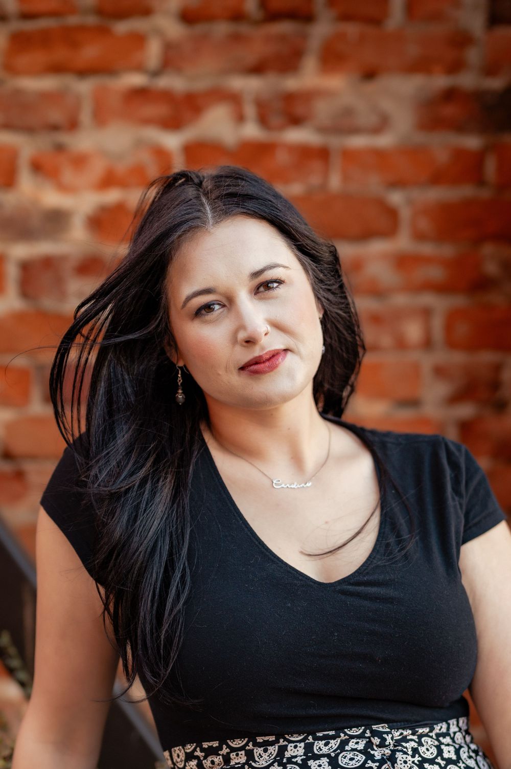 Portrait against rustic brick wall wearing black v-neck top with flowing dark hair and delicate necklace.