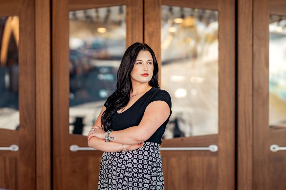 Professional portrait of a person with dark hair in a black top and patterned skirt standing with arms crossed by wooden doors.