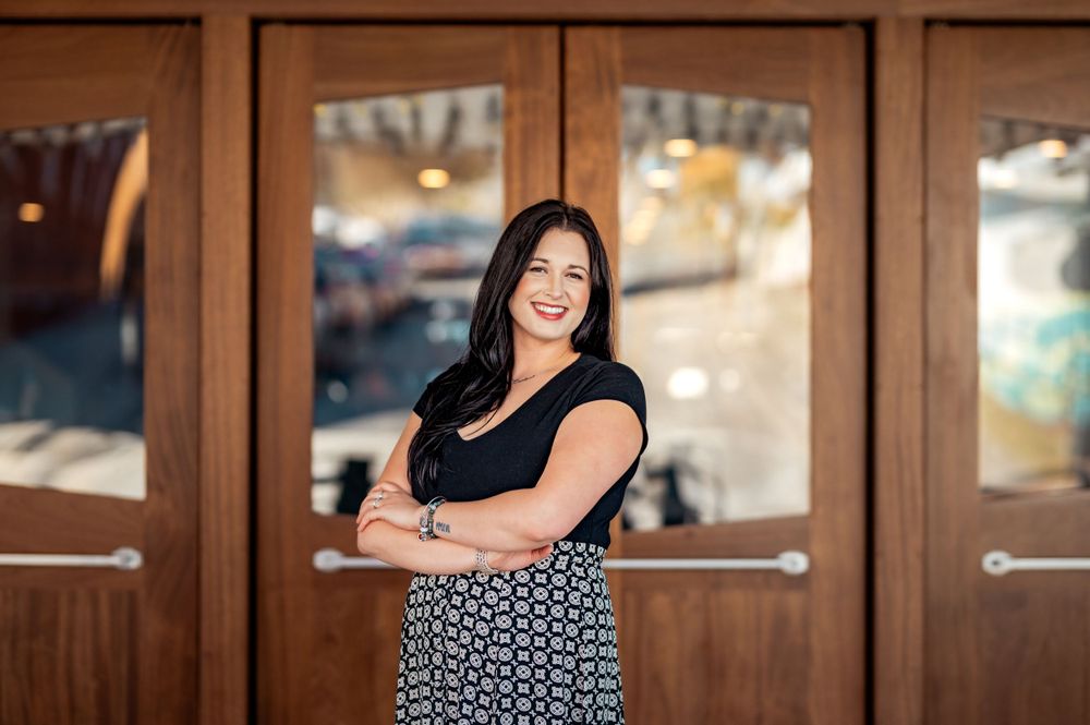 Professional portrait in front of wooden doors wearing a black top and patterned skirt with arms crossed.