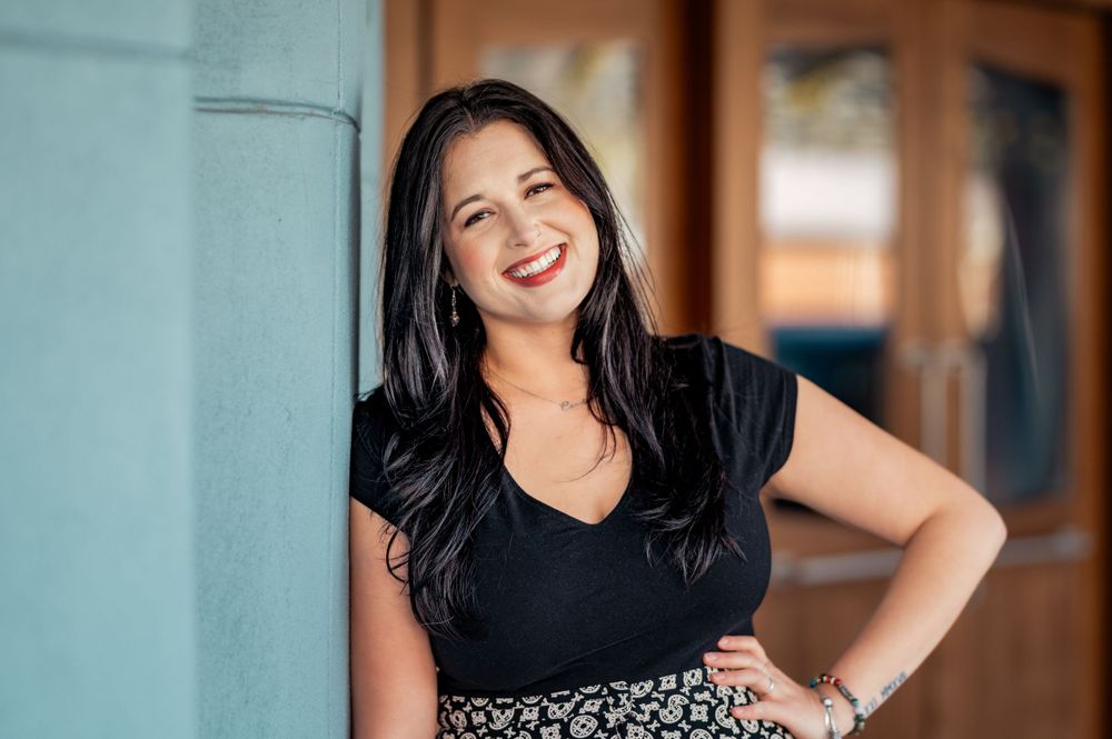 A dark-haired woman in a black top and patterned skirt smiles brightly while standing against a turquoise wall.