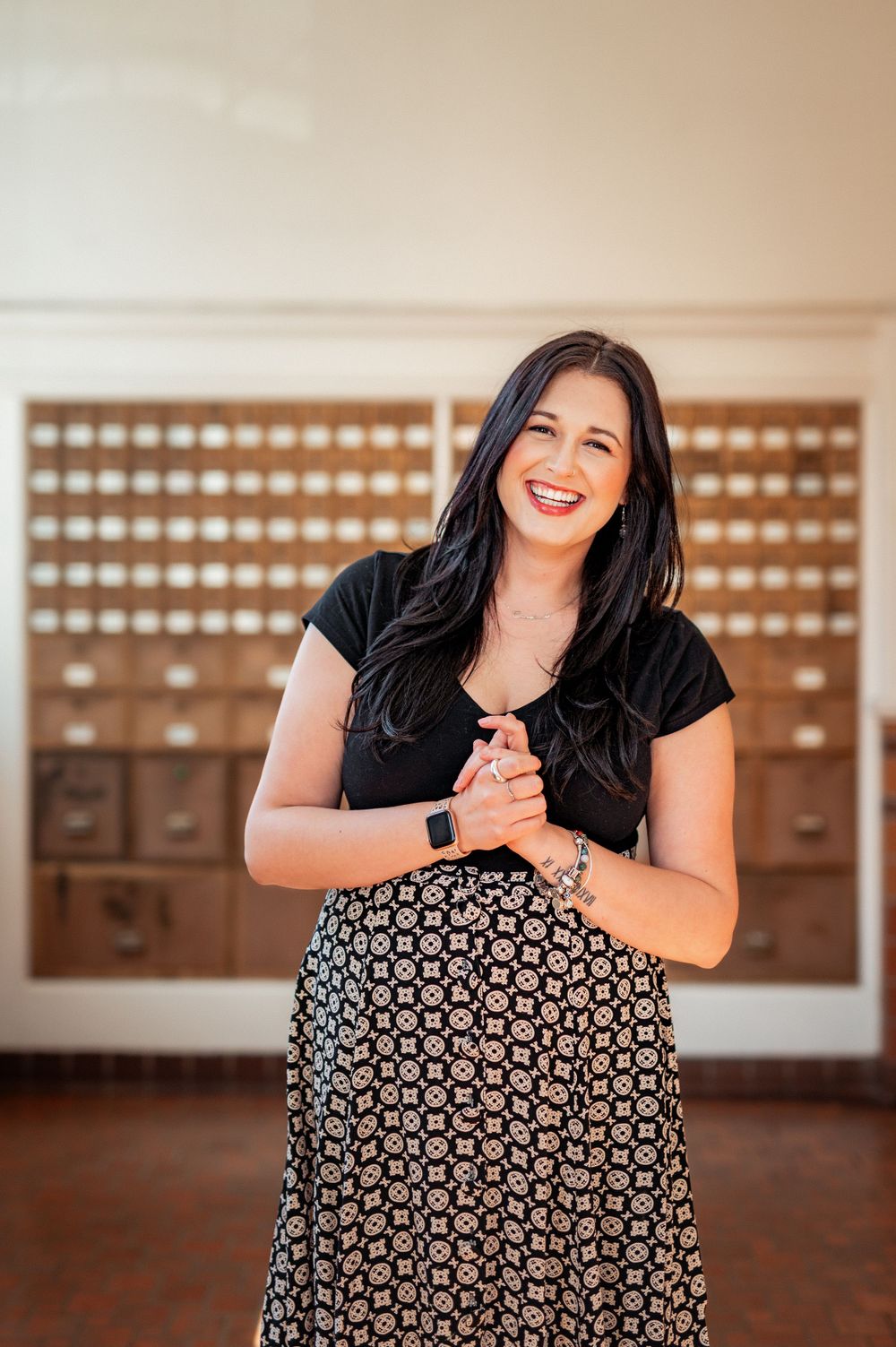 Smiling person wearing black t-shirt and patterned skirt standing in front of vintage card catalog drawers.