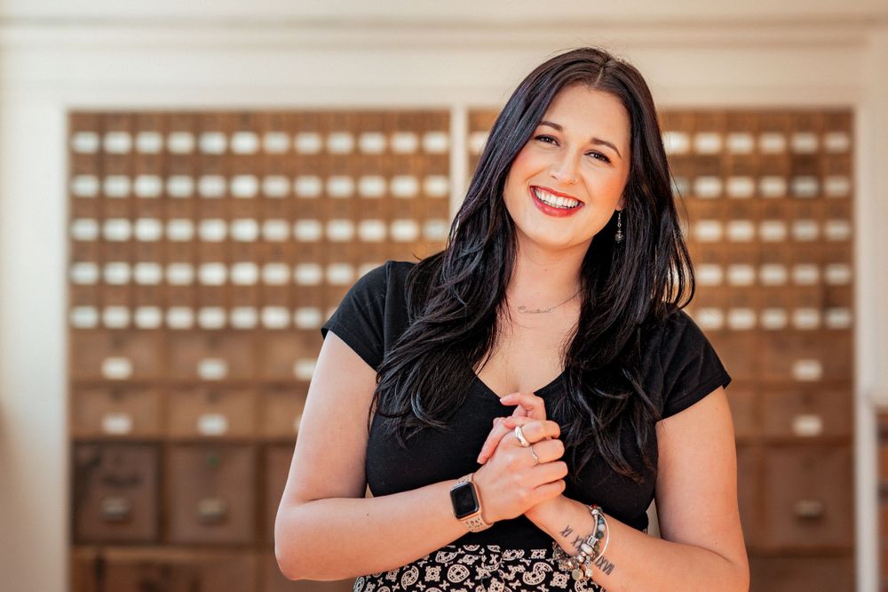 Joyous woman with long dark hair laughs while standing in front of vintage wooden card catalog drawers.