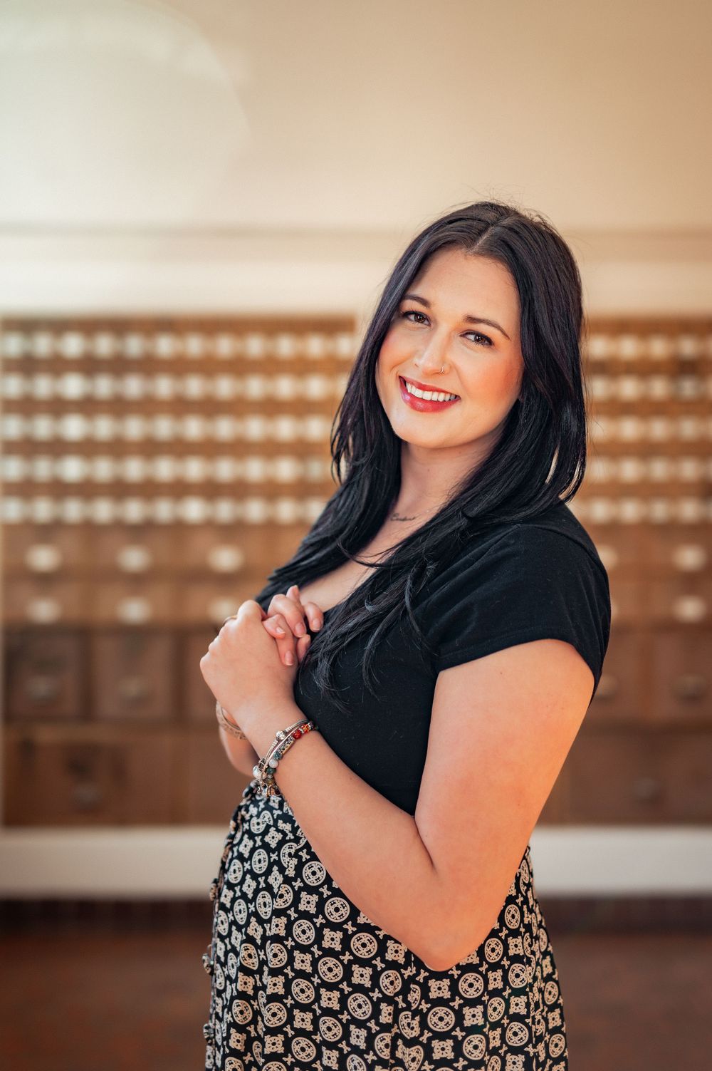 A woman smiles warmly while standing in front of vintage wooden drawers wearing a black top and patterned dress.