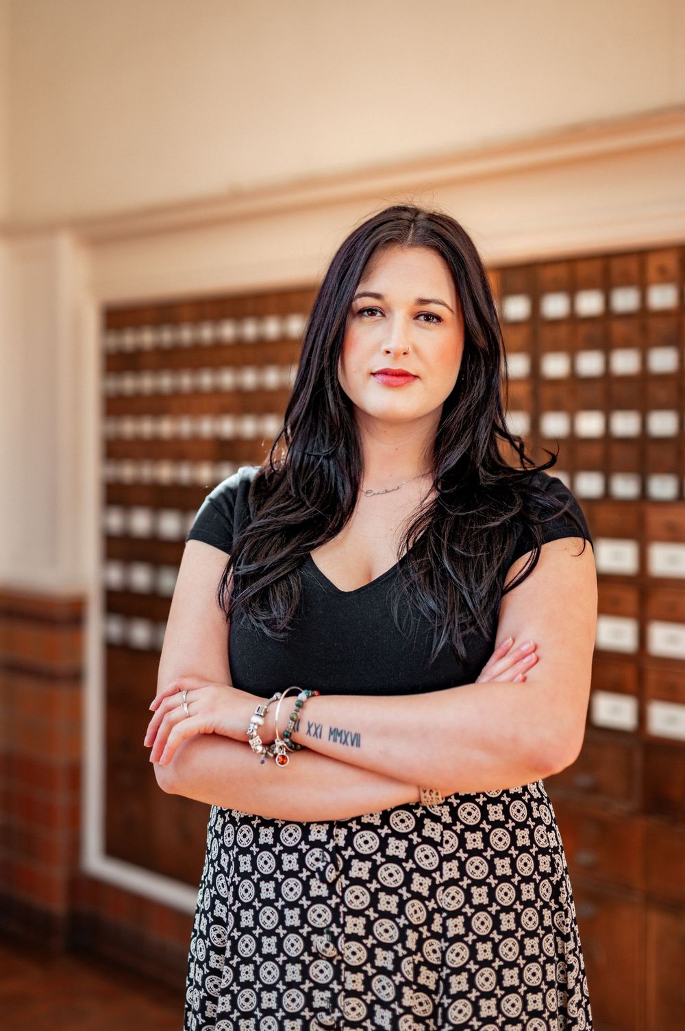 Professional portrait of someone with long dark hair wearing a black top and patterned skirt standing with arms crossed.