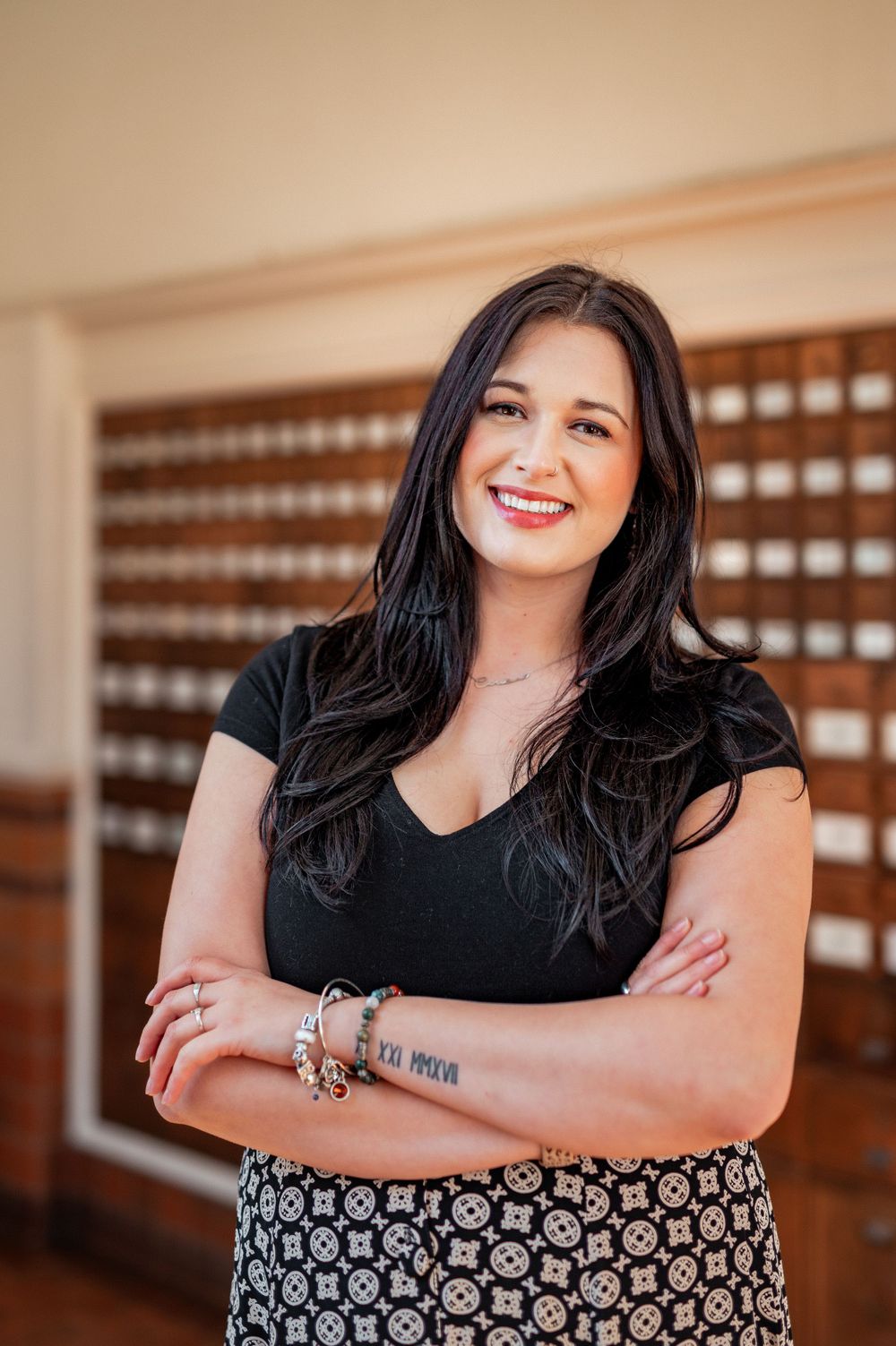 Professional portrait of someone with dark hair in a black top and patterned skirt standing with crossed arms in an office.